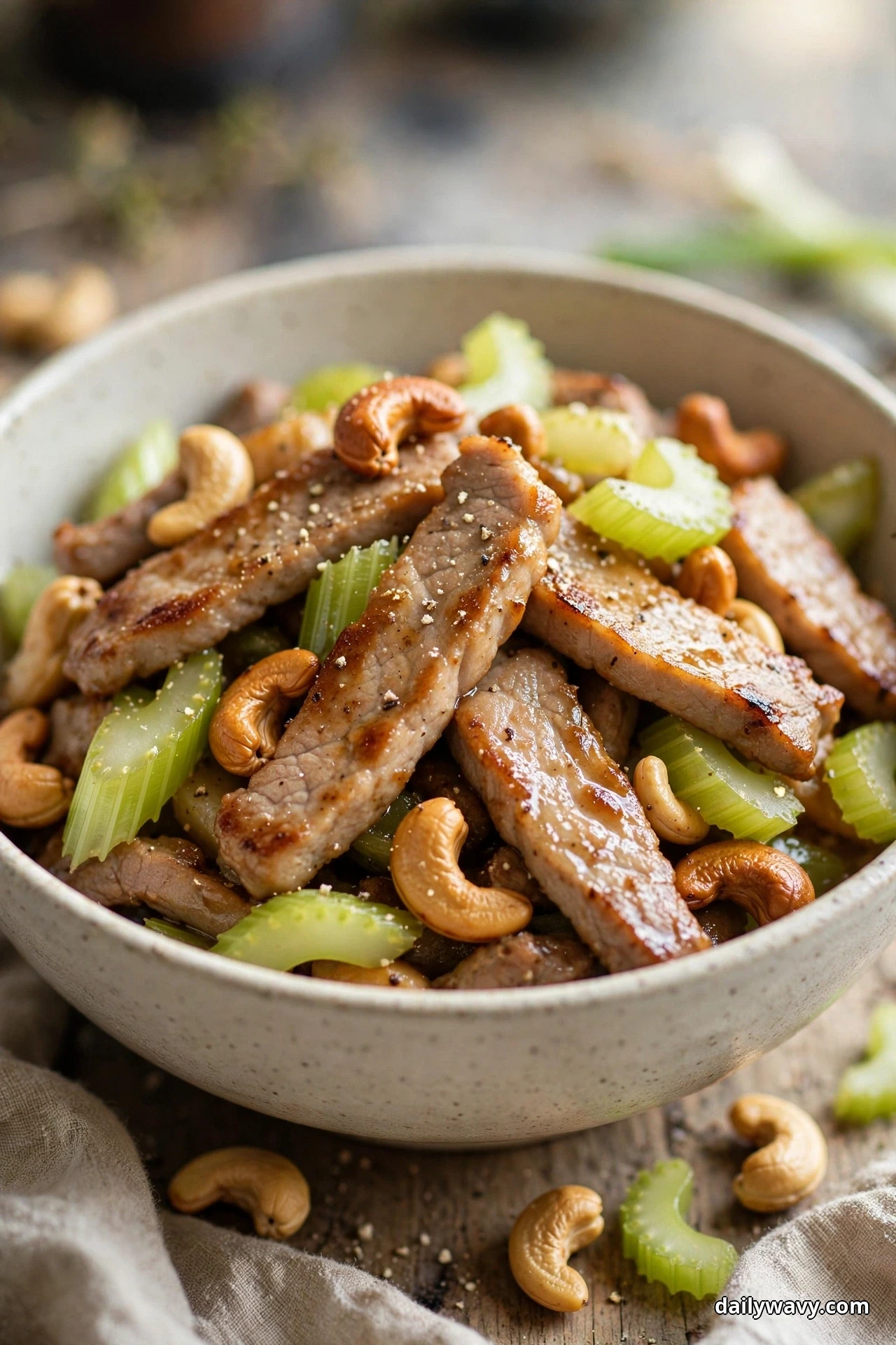 A ceramic bowl of seared pork strips, diagonal celery slices and roasted cashews sits on a wooden table with loose cashews and celery scattered beside it