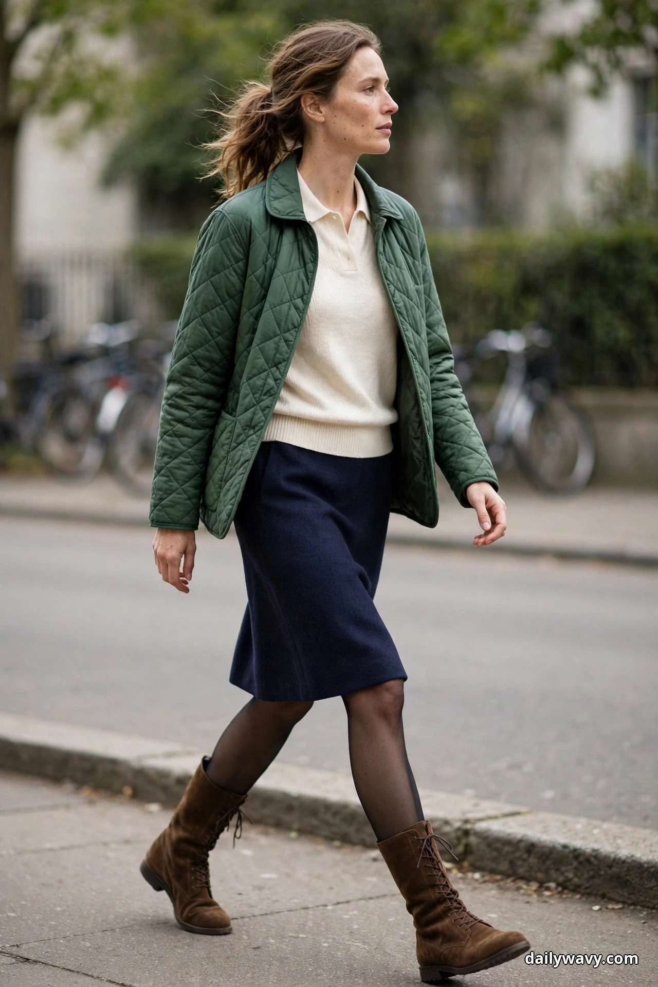 A woman wearing a forest-green diamond-quilted jacket over a cream polo knit with a navy A-line skirt, sheer tights and brown suede lace-up mid-calf boots walks along a city sidewalk, shown full length
