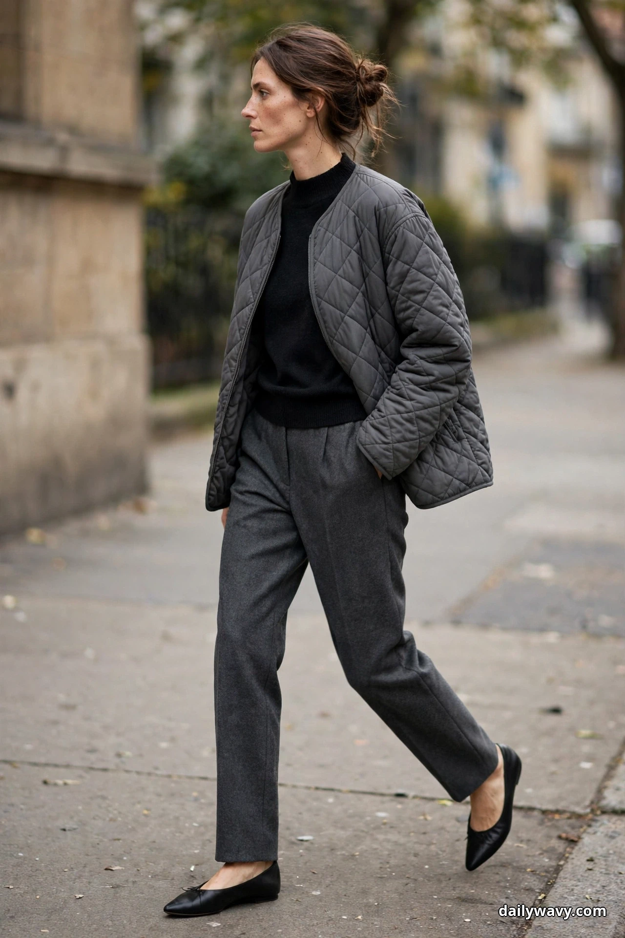 A woman wearing a charcoal diamond-quilted jacket over a black mock-neck knit with charcoal wool trousers and black pointed flats walks along a city sidewalk, shown full length