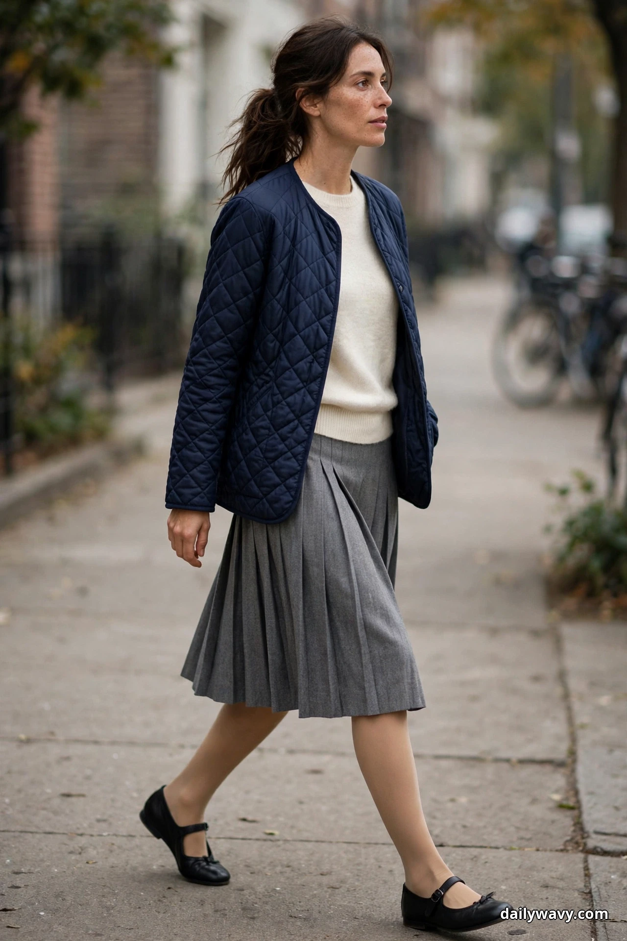A woman wearing a navy diamond-quilted jacket over a cream light sweater with a gray pleated skirt and black Mary Jane flats walks along a city sidewalk, shown full length