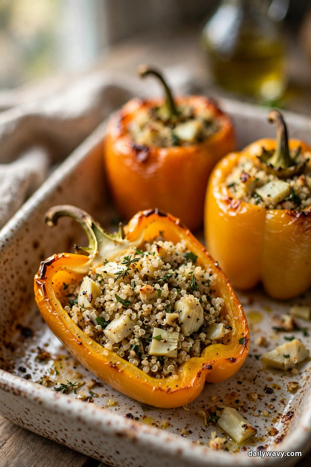 Yellow and orange bell peppers stuffed with quinoa and artichoke heart pieces sit in a speckled baking dish, one pepper halved to show the filling inside