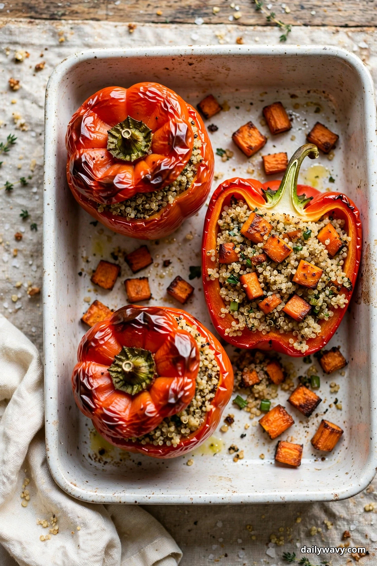 Seen from above, red bell peppers filled with quinoa and caramelized roasted carrot cubes rest in a white speckled baking dish, one pepper halved to show the filling