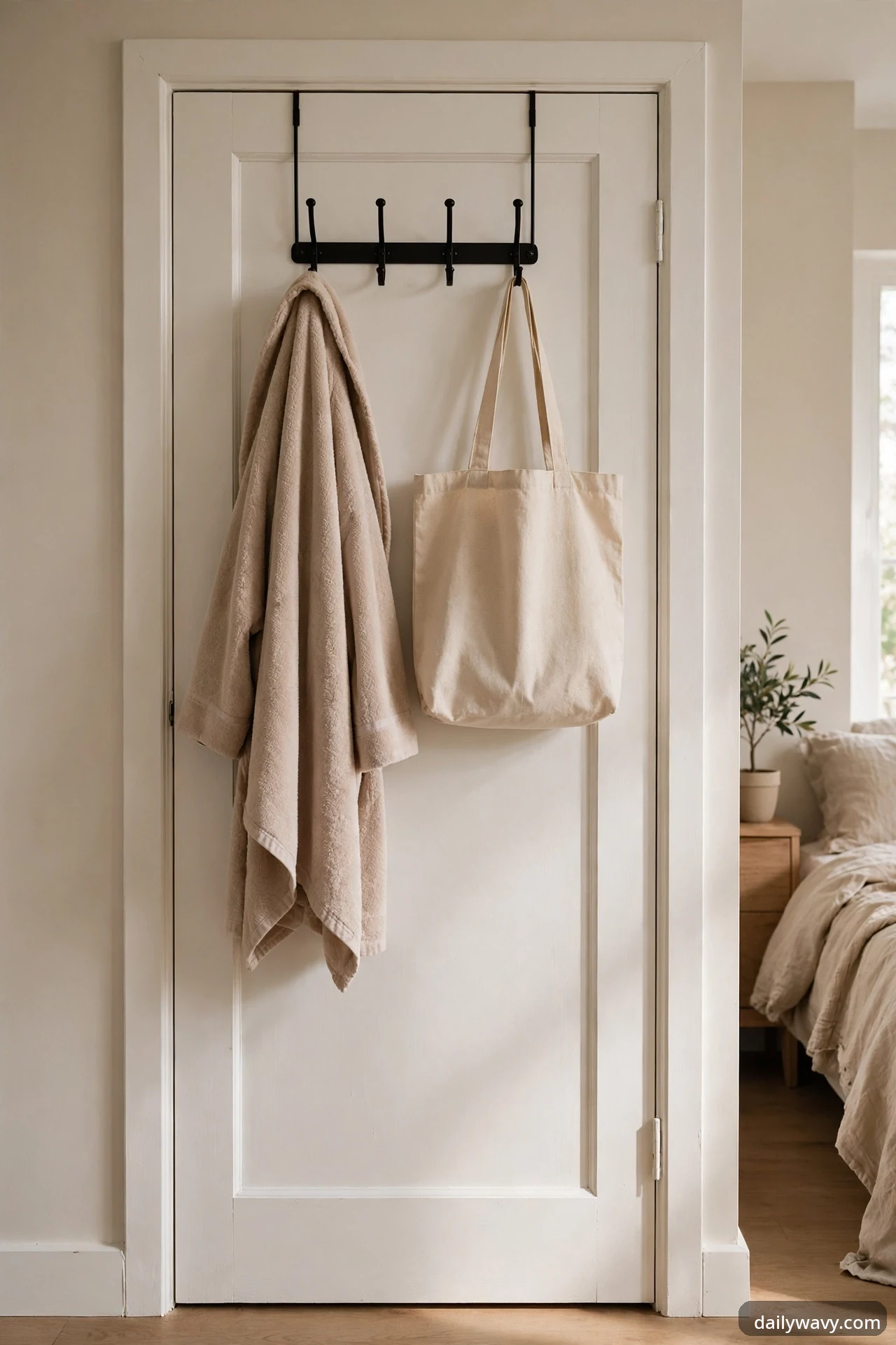 A black over-the-door hook rack holding a beige bathrobe and a canvas tote bag on a white bedroom door, with the bed visible through the doorway.