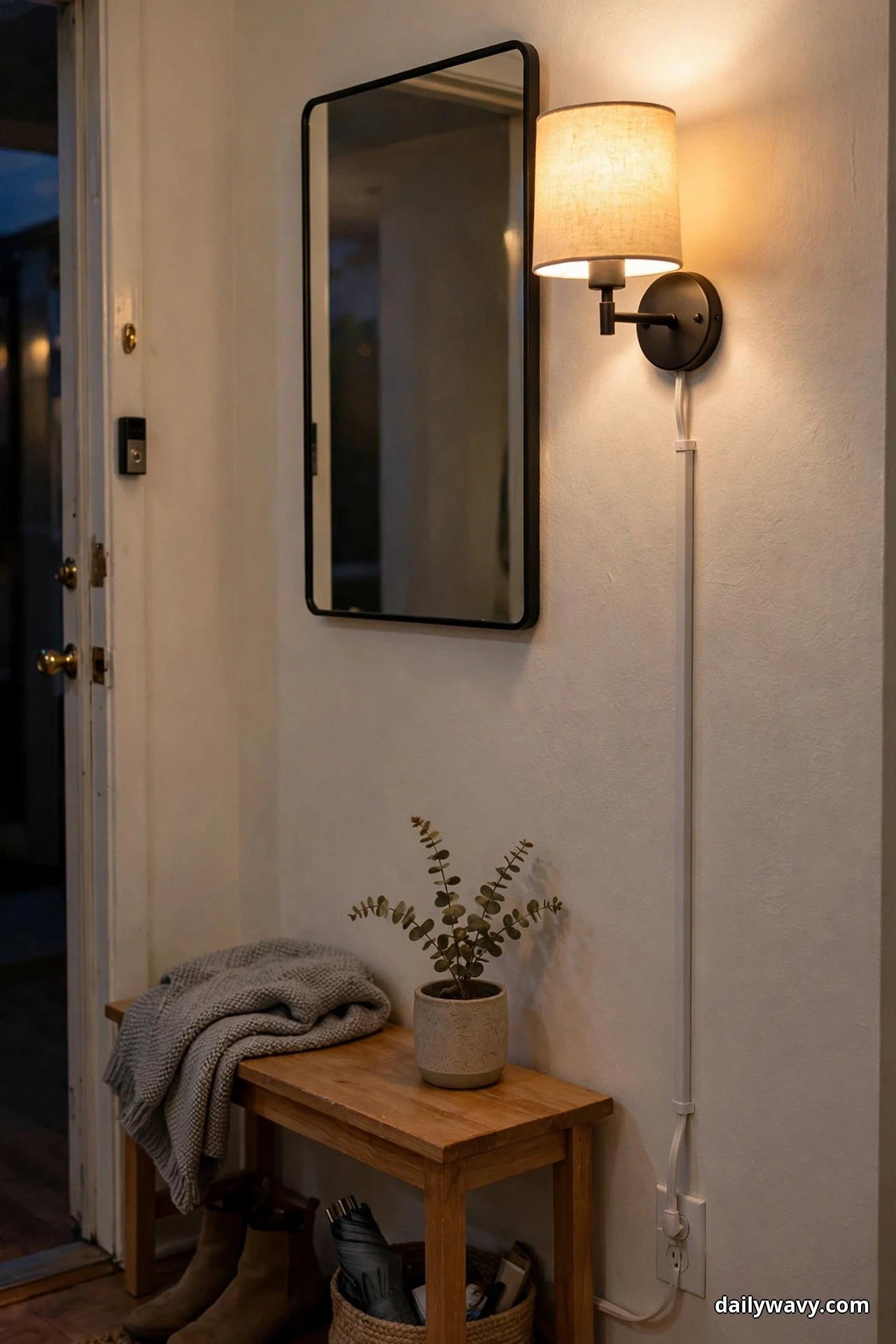 A plug-in wall sconce with a linen shade glows warmly beside a black-framed mirror, its white cord cover running straight down the wall to the outlet