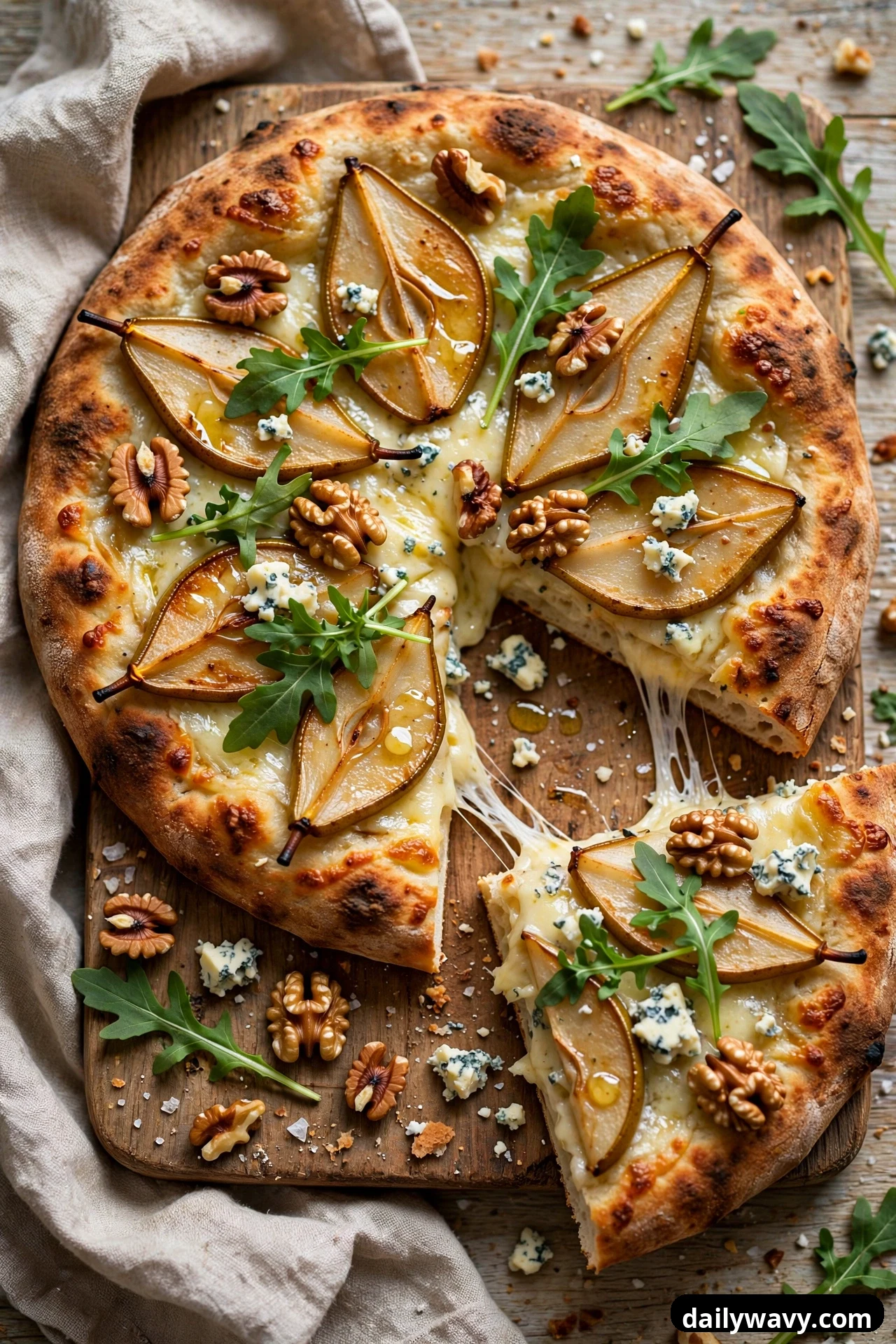 A flatbread topped with roasted pear slices, walnut halves, crumbled blue cheese, and fresh arugula leaves sits on a wooden board with one slice pulled away.