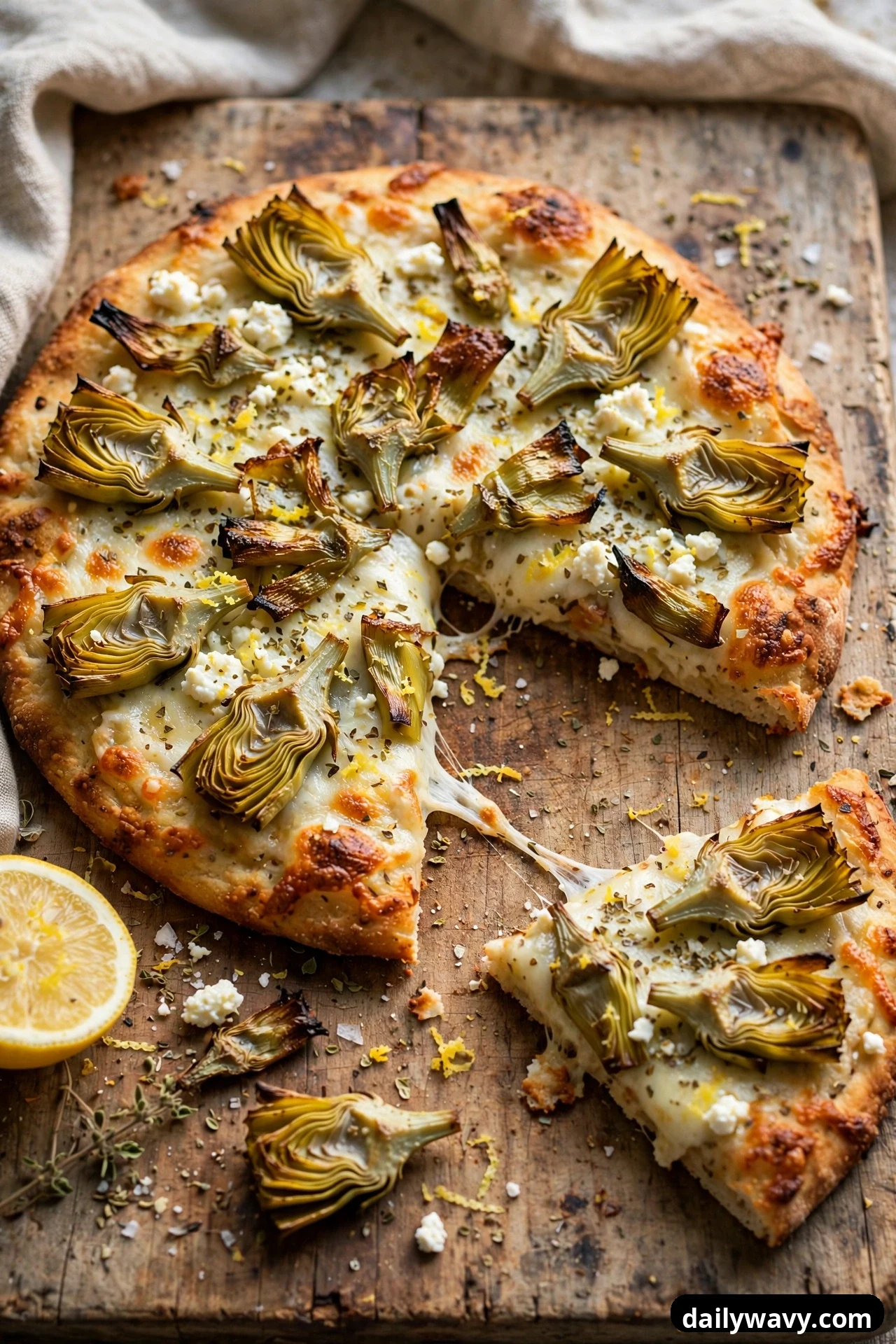A round flatbread topped with crispy roasted artichoke heart halves, melted mozzarella, crumbled feta, dried oregano, and lemon zest sits on a wooden board with a lemon half beside it.