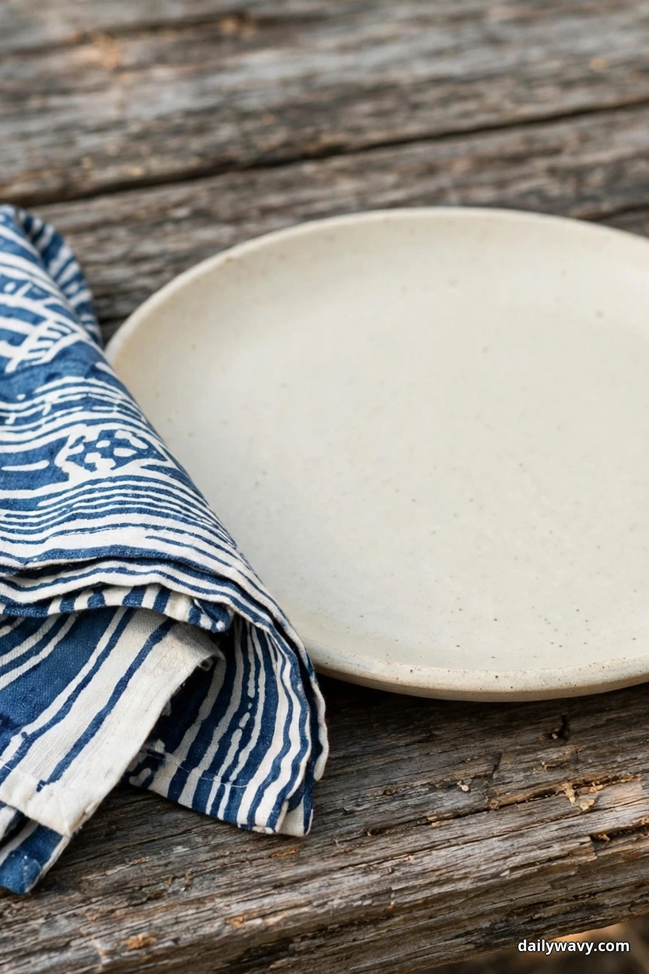 A folded indigo block-printed napkin rests beside a plain cream stoneware plate on a weathered wood table