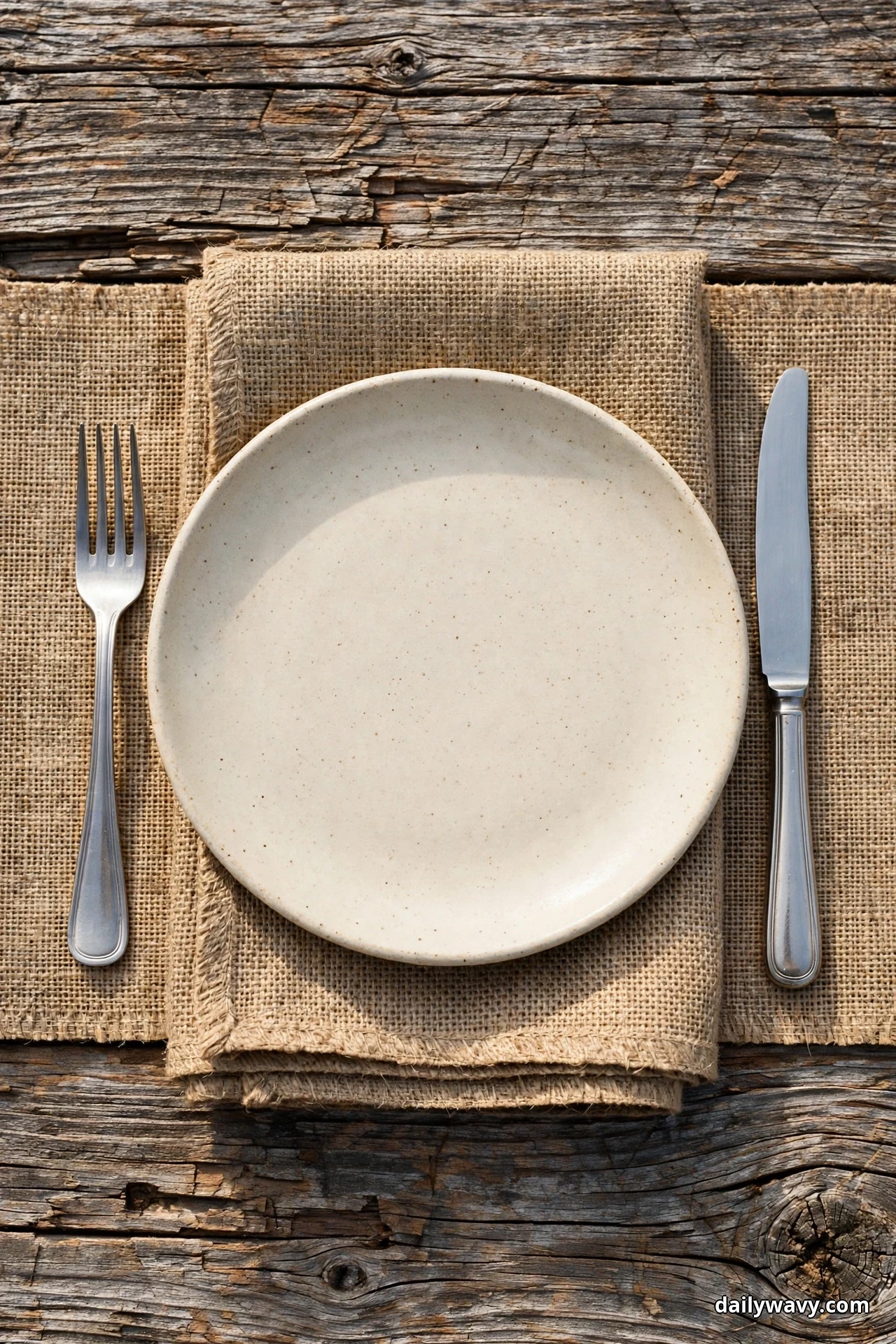 An overhead view shows a cream stoneware plate centered on a folded burlap fabric strip with a fork at the left and a knife at the right on weathered wood