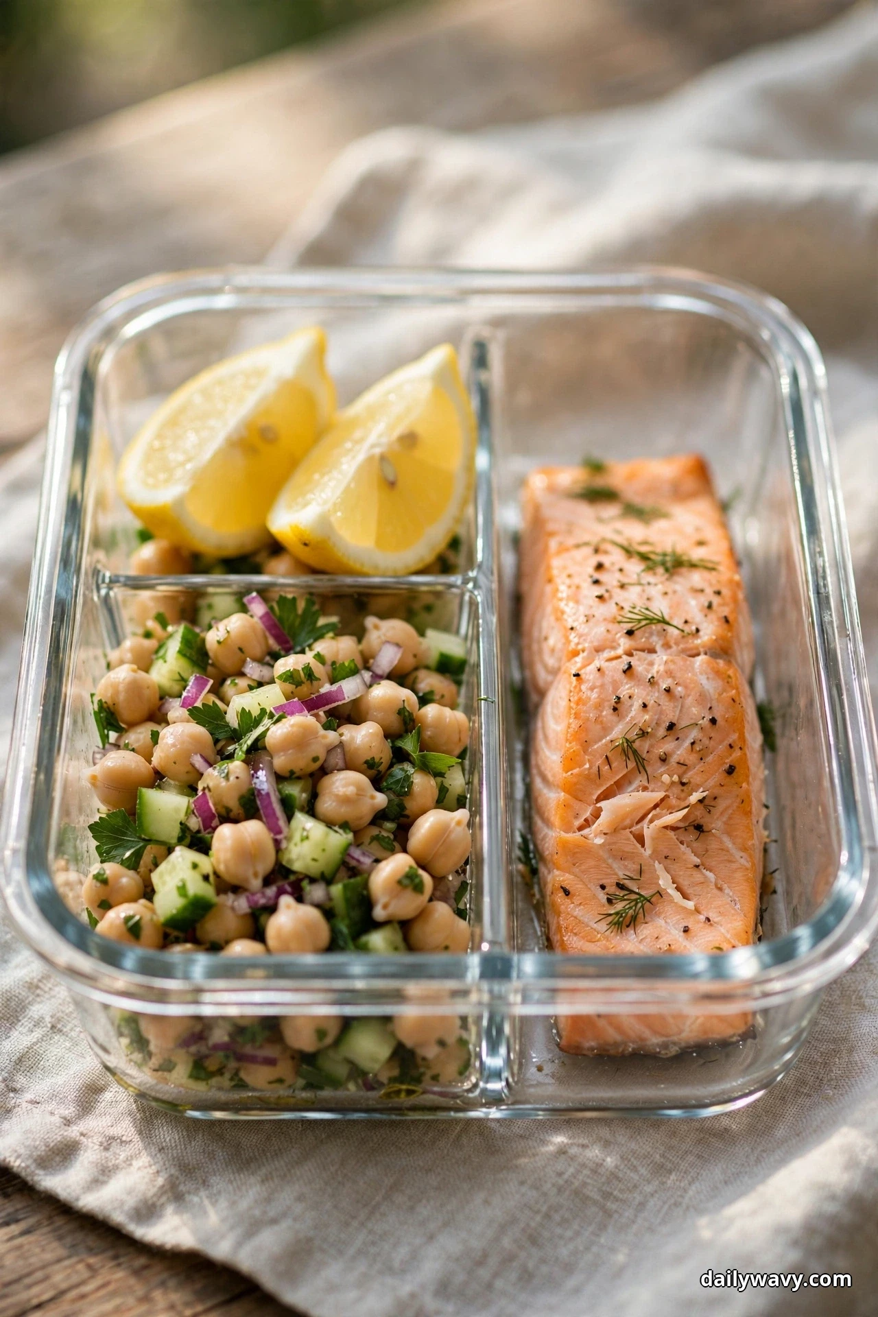 A divided glass container holds a chickpea salad with cucumber and red onion, two lemon wedges and a baked salmon fillet