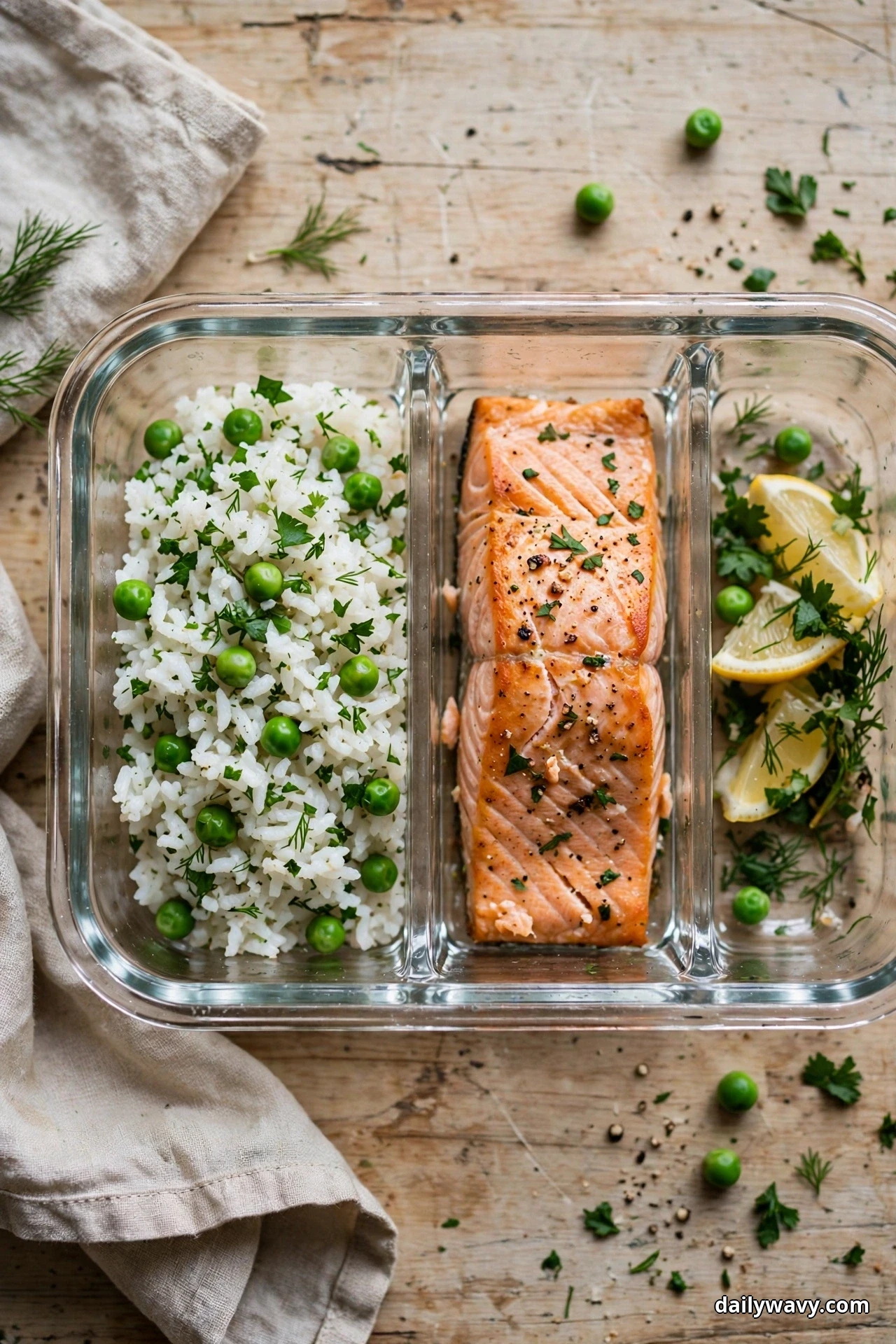 An overhead view shows a divided glass container holding white rice studded with peas and herbs, a baked salmon fillet, and lemon slices with herbs in the third compartment
