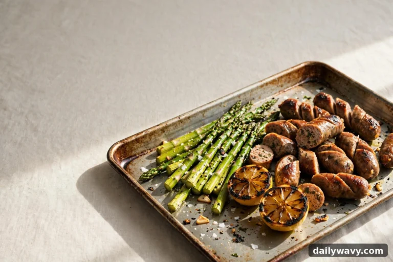 An angled view of a sheet pan shows sliced sausage pieces and browned links piled beside roasted asparagus spears and two charred lemon halves.