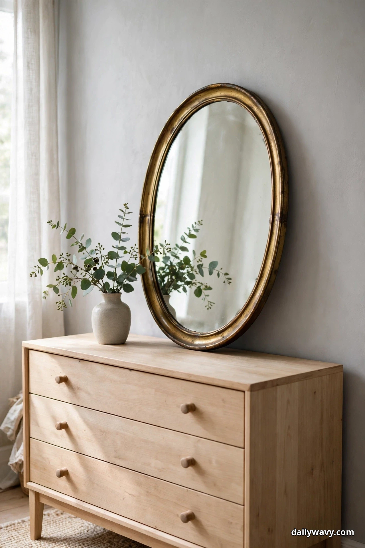 A large oval vintage mirror with a brass frame leans on a light wood dresser against a pale gray wall, with a small vase of eucalyptus beside it