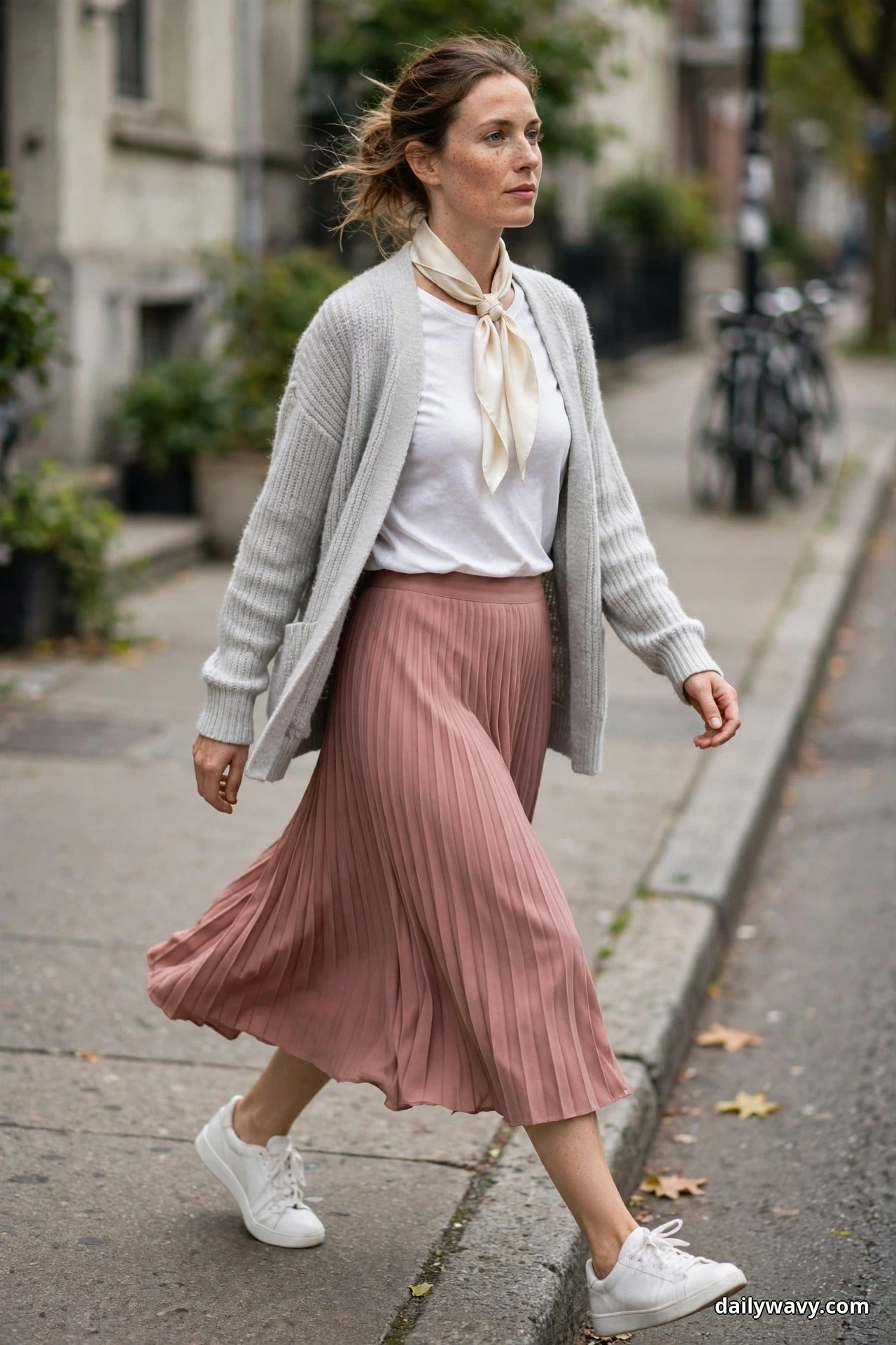 A woman walks along a city sidewalk wearing a light-gray open cardigan over a white tee with a dusty-rose pleated midi skirt, a cream skinny scarf tied at her neck and white sneakers