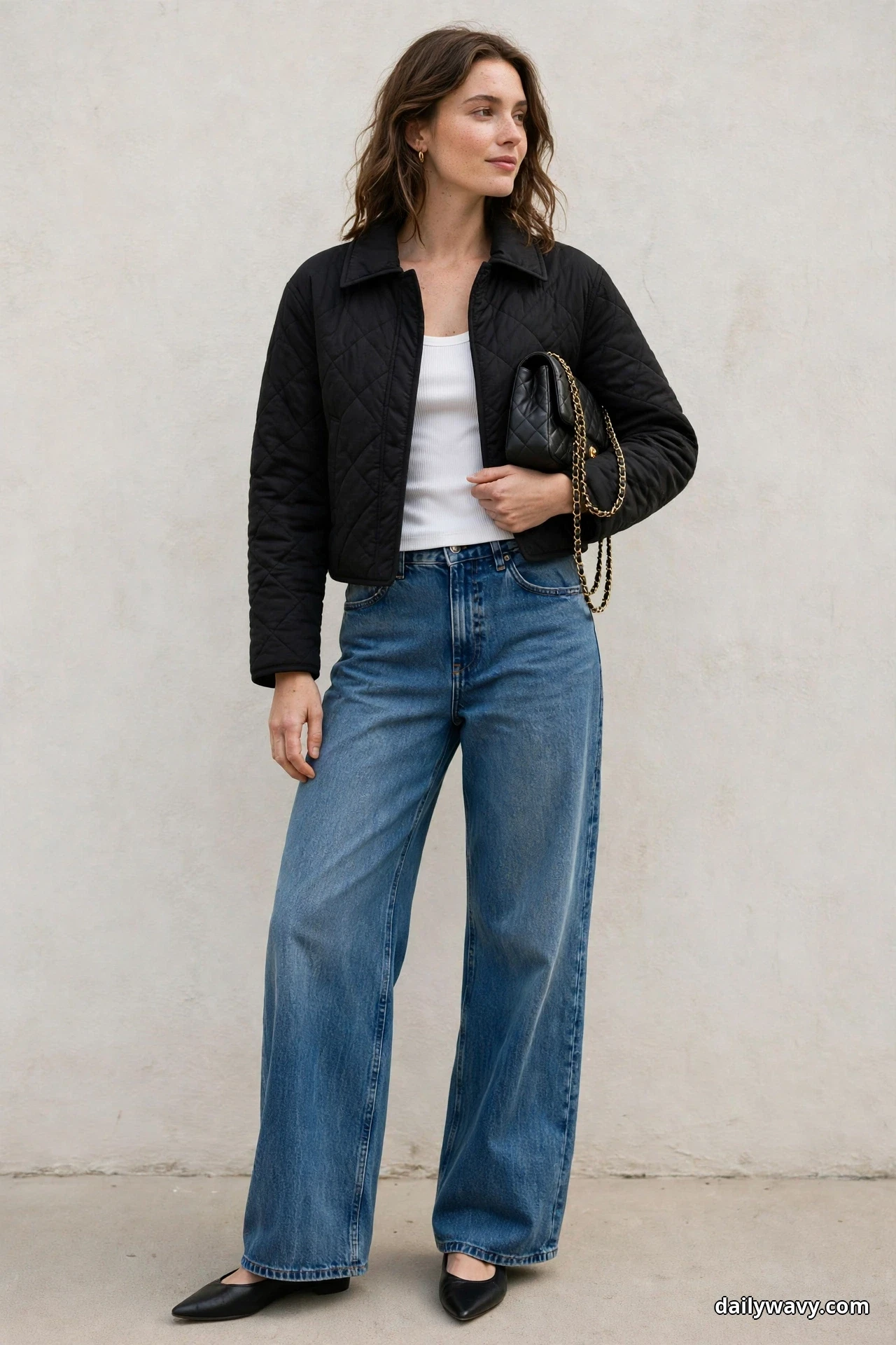 A woman wearing wide-leg jeans, a white tank and a cropped black quilted jacket stands full length against a plain wall holding a small black quilted shoulder bag with a gold chain strap in her hand against her side, with black pointed flats below the hems