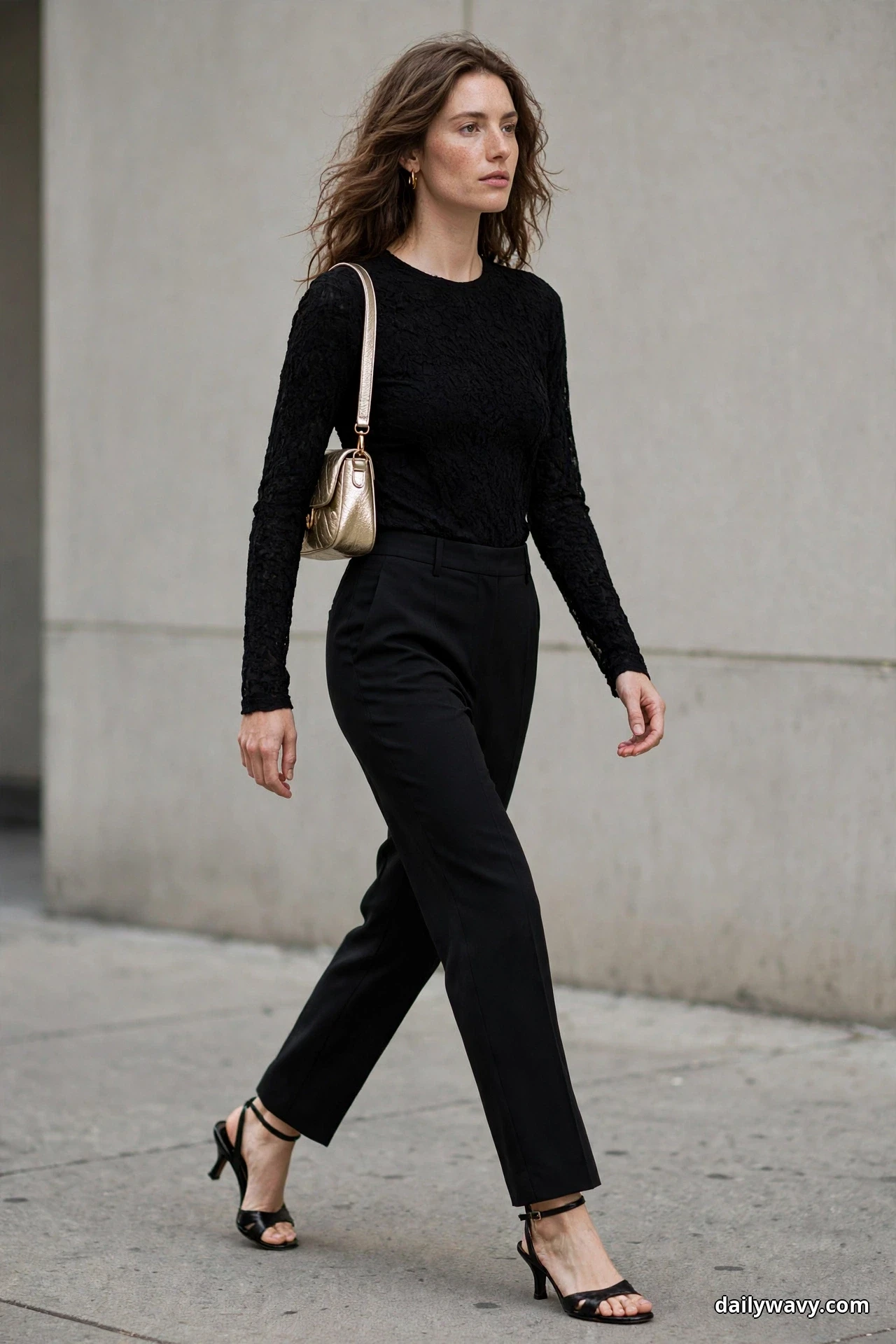 A woman wearing straight black trousers, a black long-sleeve lace top and black heeled sandals walks along a sidewalk with a small metallic champagne shoulder bag on her shoulder at her hip