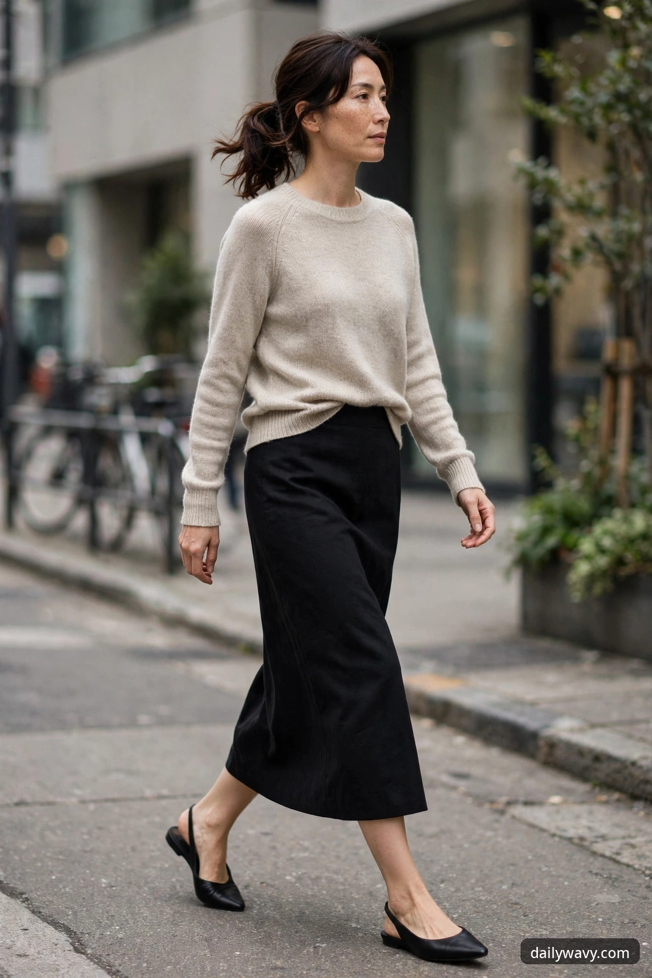 A woman walks on a city street wearing an oatmeal knit sweater tucked into a straight black midi skirt with black pointed slingback flats.