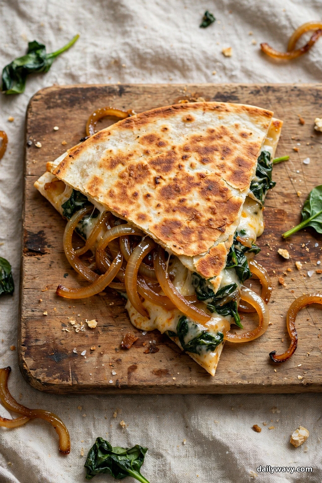An overhead view shows a golden folded quesadilla on a wooden board with strands of caramelized onion and melted cheese spilling from its spinach filling