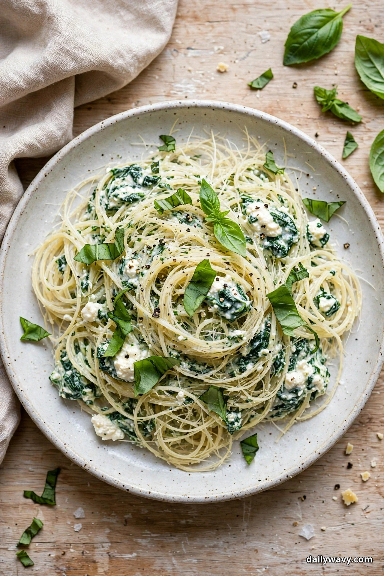 Seen from above, a speckled shallow bowl of angel hair pasta tangled with wilted spinach, ricotta and torn basil leaves sits on a wooden table with linen beside it