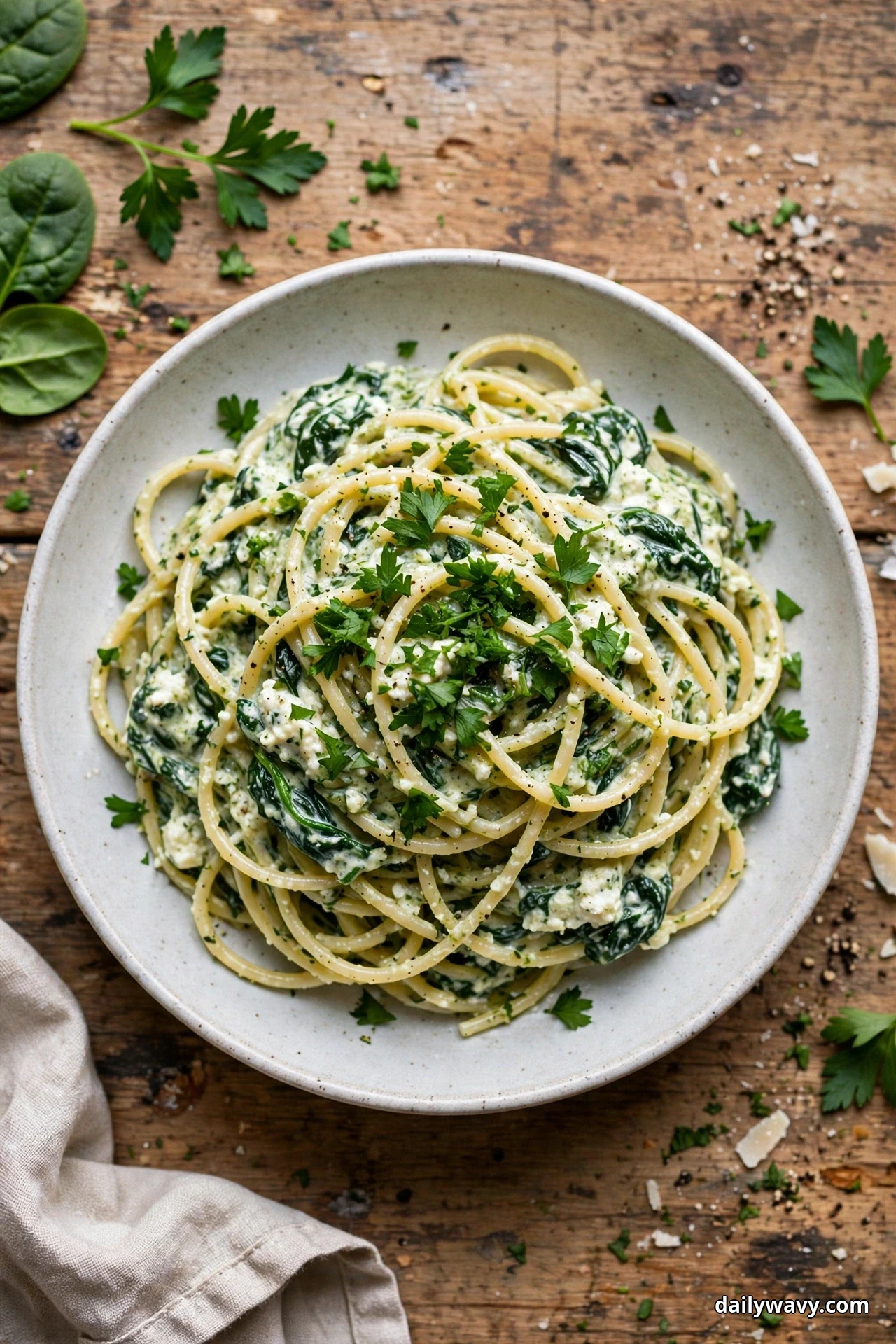 Seen from above, a speckled bowl of spaghetti coated in creamy spinach ricotta sauce and topped with chopped flat-leaf parsley sits on a rustic wooden table