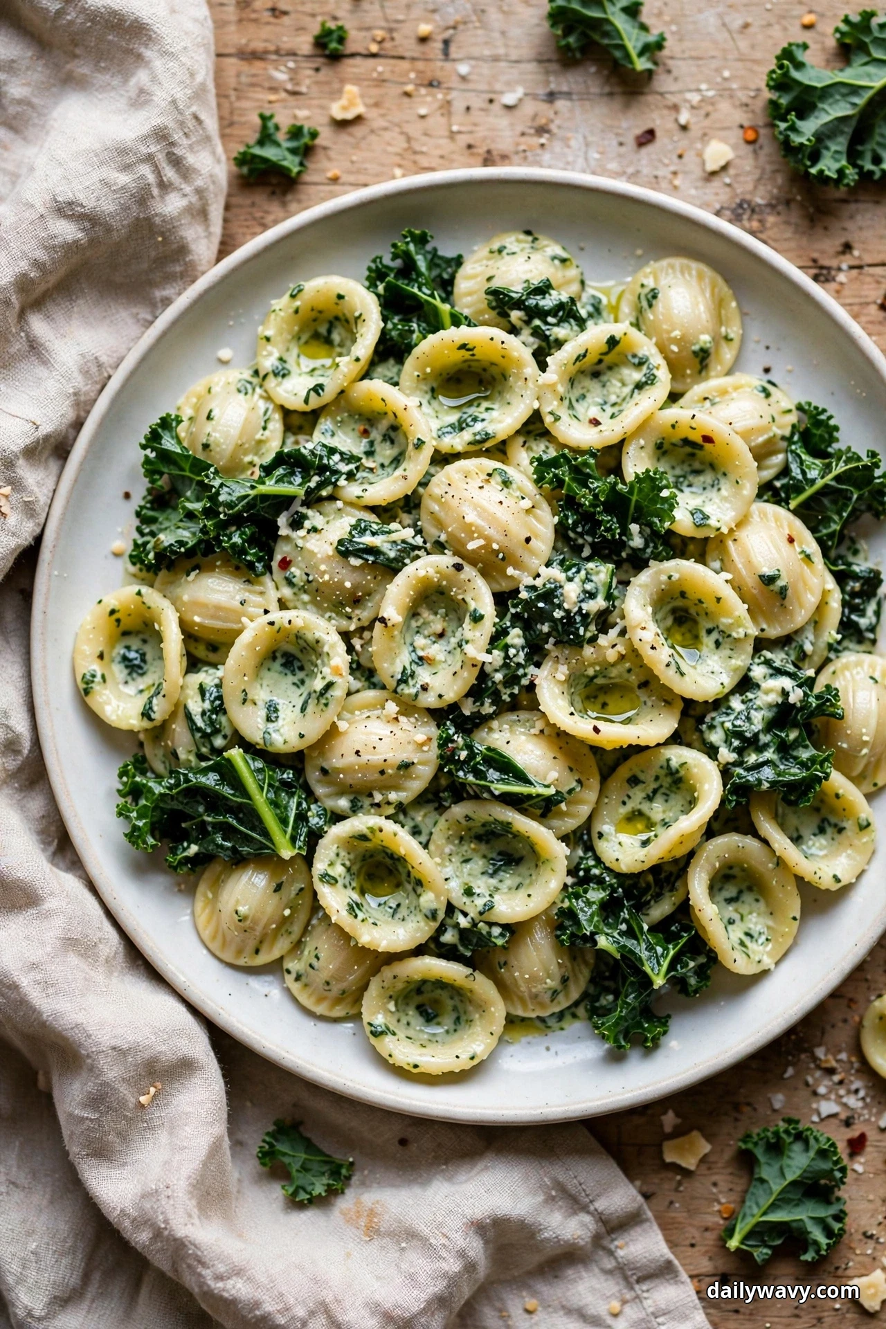 Seen from above, a plate of orecchiette cups holding creamy ricotta sauce and mixed with dark kale leaves sits on a wooden table with linen and loose kale leaves around it