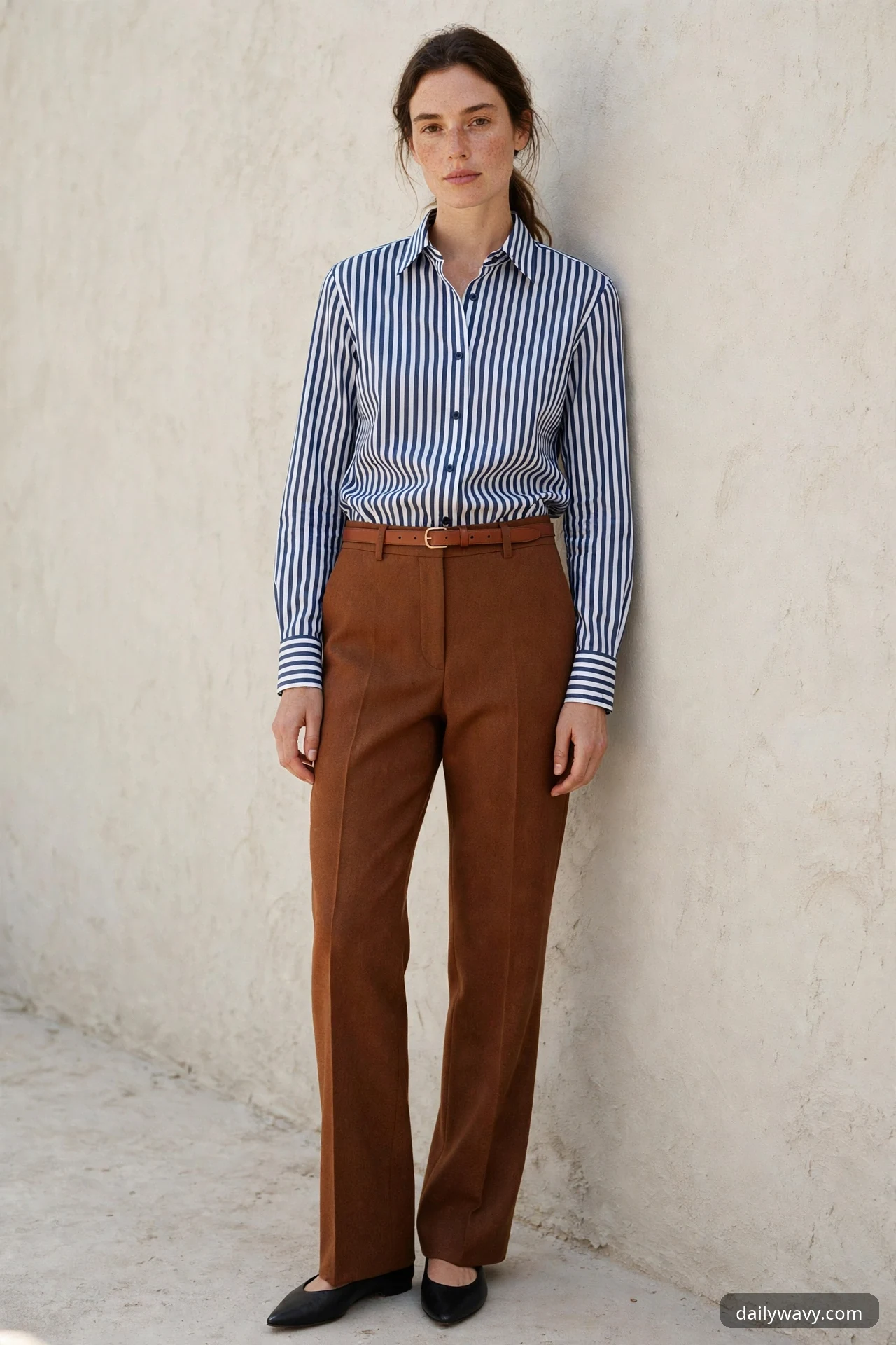 A woman in a blue-and-white striped shirt tucked into rust-brown high-rise trousers with a slim tan belt, wearing black pointed flats, standing against a beige wall.