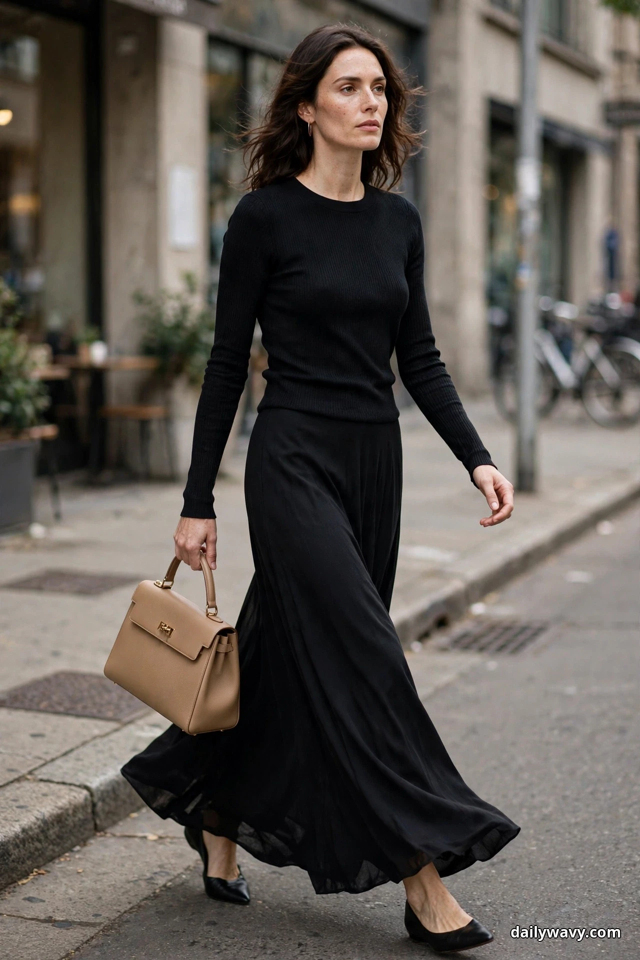 A woman walks along a city street wearing a fitted black long-sleeve ribbed knit with a flowing black maxi skirt, holding a tan structured top-handle bag, with black pointed flats