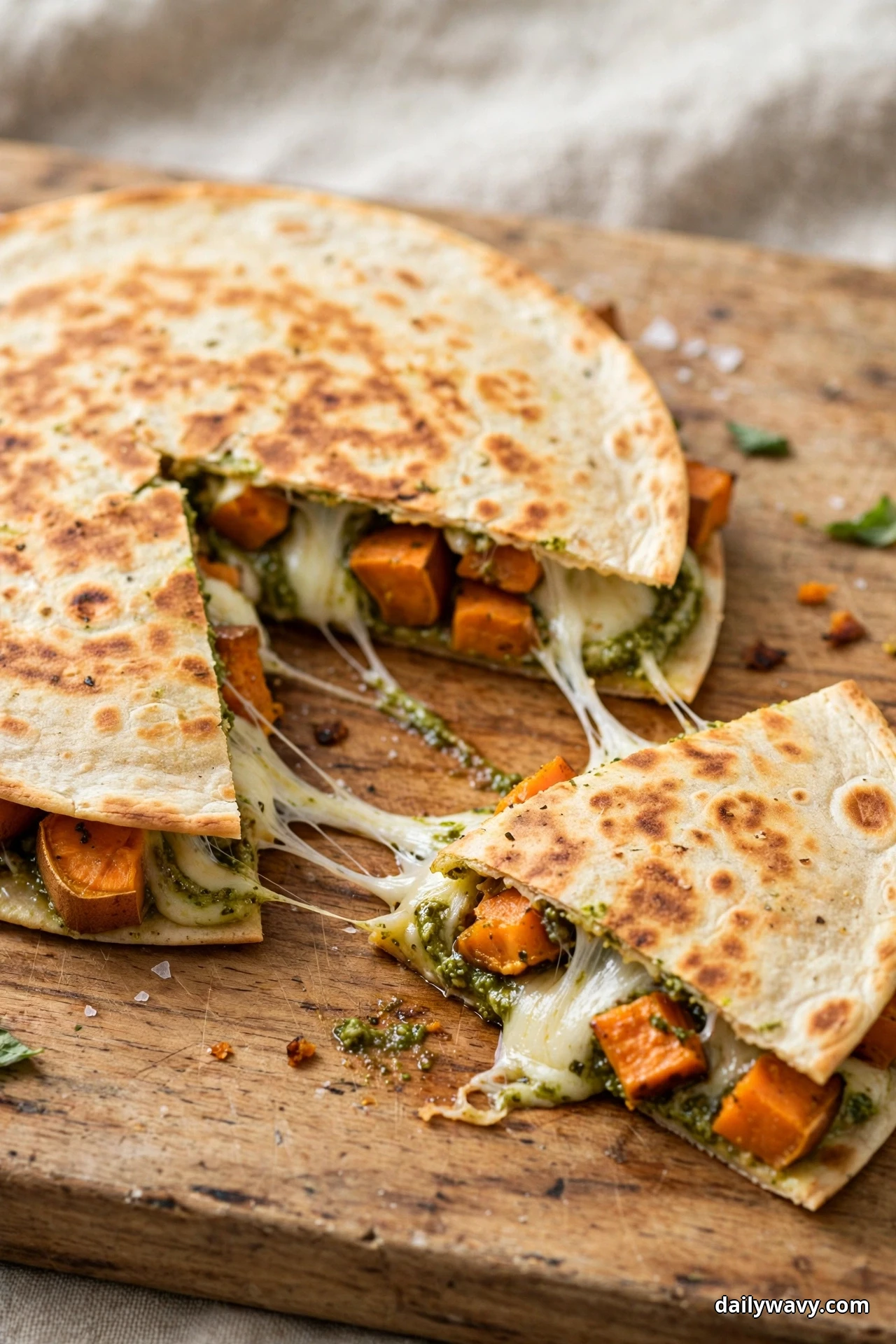 A round sweet potato quesadilla cut into wedges on a wooden board has one wedge pulled away with a cheese pull, showing green pesto and sweet potato cubes in the filling