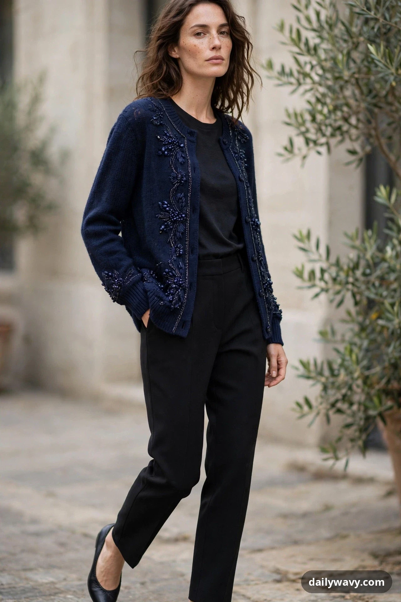 A woman walks outdoors wearing a navy beaded cardigan open over a black top, paired with black tapered trousers and black flats.