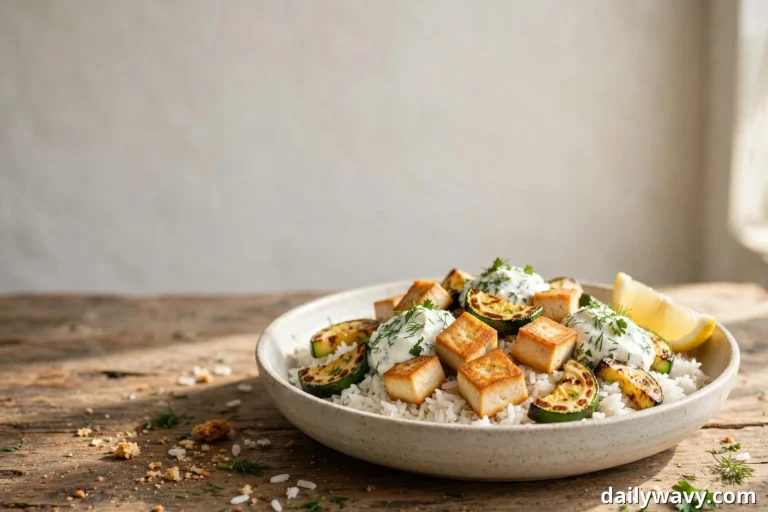 A wide view of a tofu rice bowl shows golden seared tofu cubes, charred zucchini half-moons, and dollops of herbed yogurt over white rice in a shallow speckled bowl, with a lemon wedge resting at the rim
