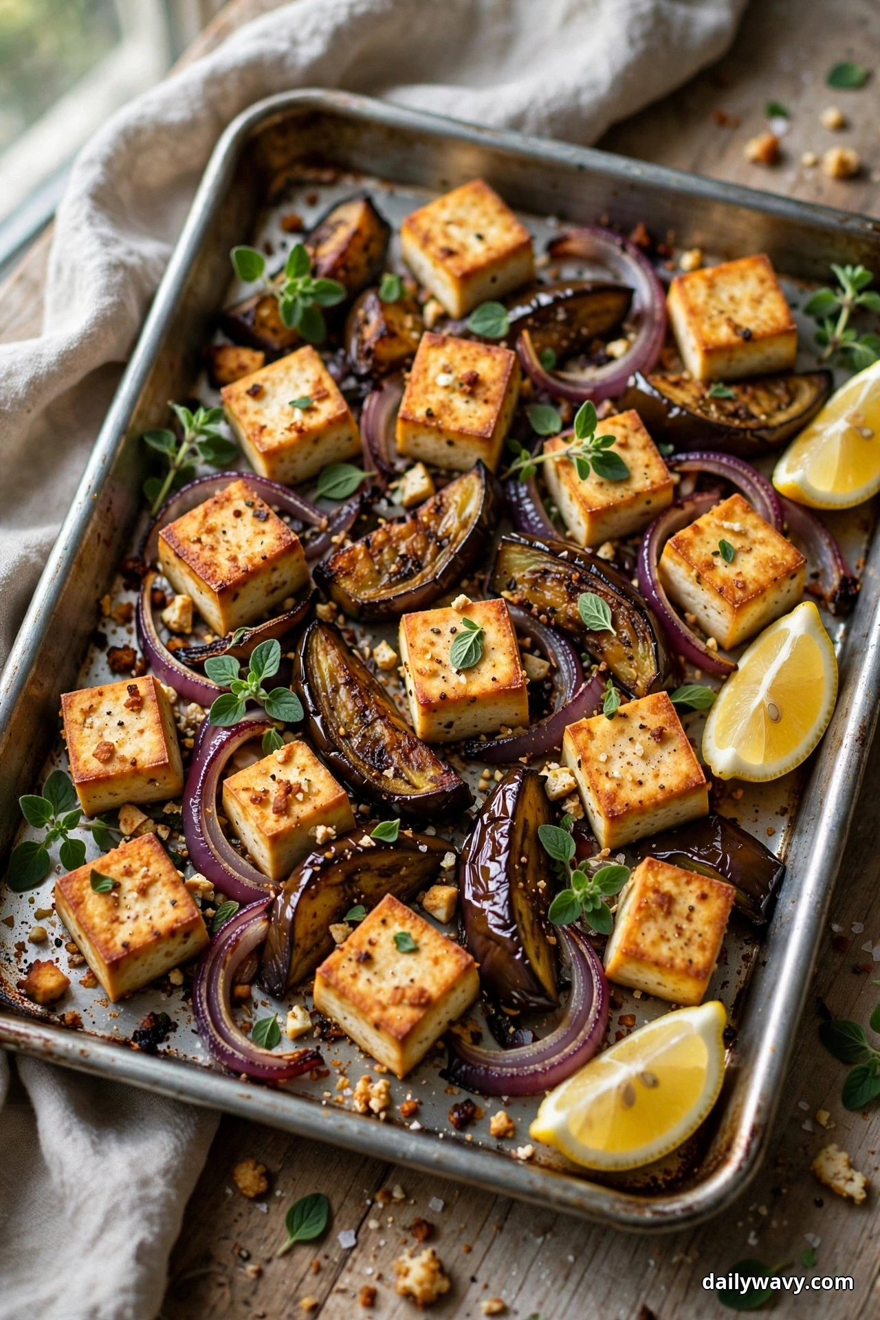 Golden roasted tofu cubes, caramelized eggplant wedges and red onion rings fill a sheet pan, finished with fresh oregano leaves and lemon wedges