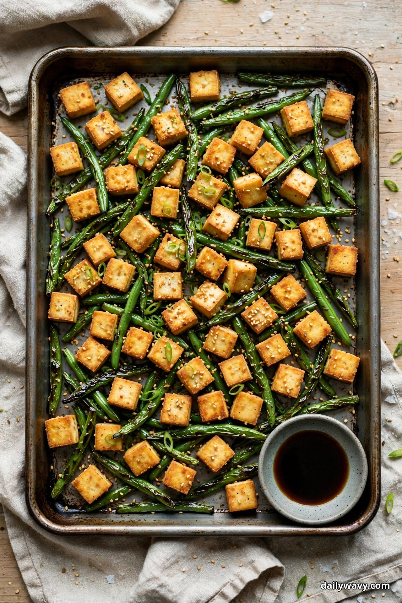 Seen from above, a sheet pan of golden tofu cubes and blistered green beans is sprinkled with sesame seeds and sliced scallions, with a small bowl of soy sauce for dipping