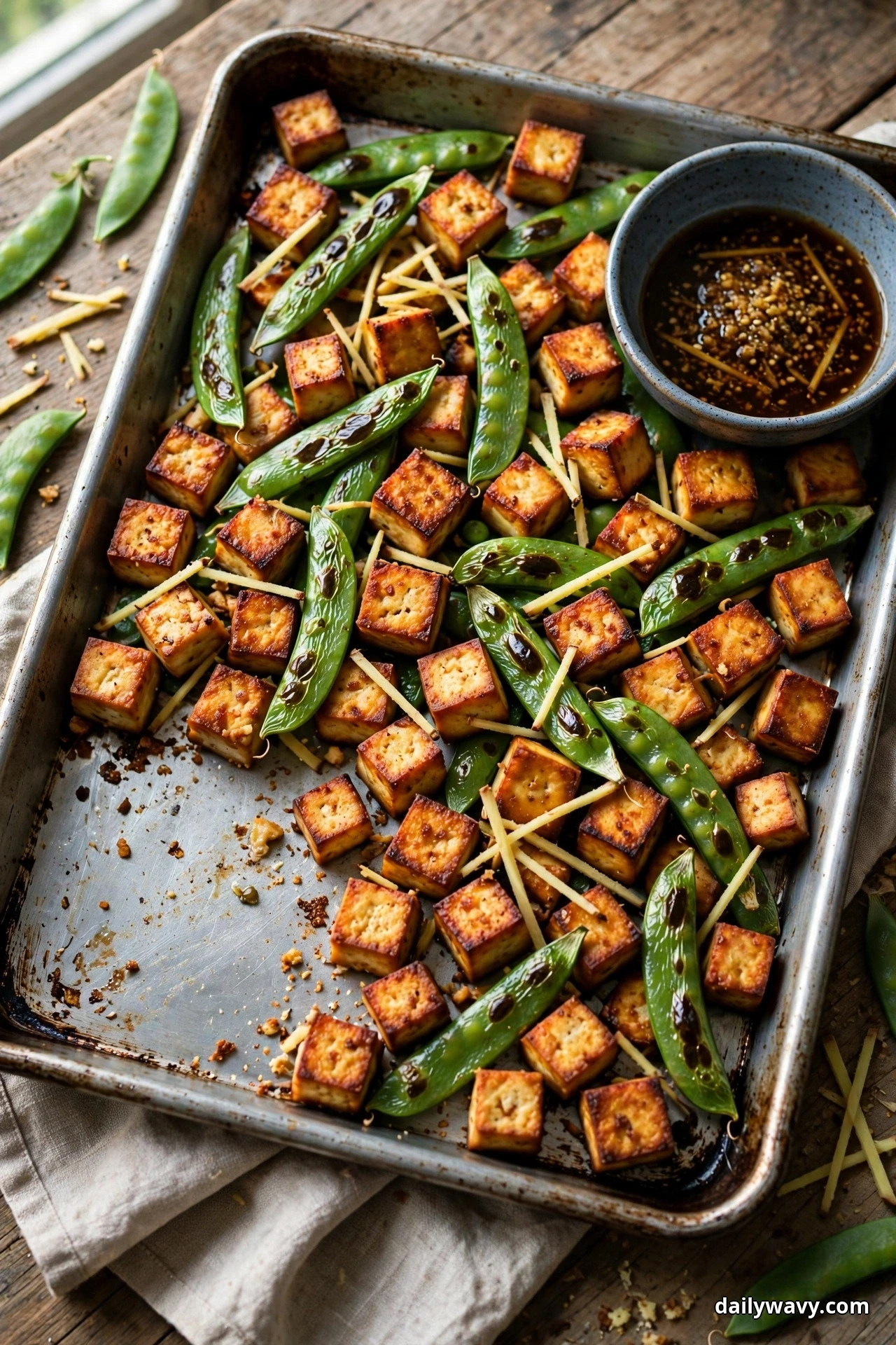 A sheet pan with one serving removed holds golden tofu cubes and bright green sugar snap peas scattered with thin strips of fresh ginger, with a bowl of dark tamari dip at the corner