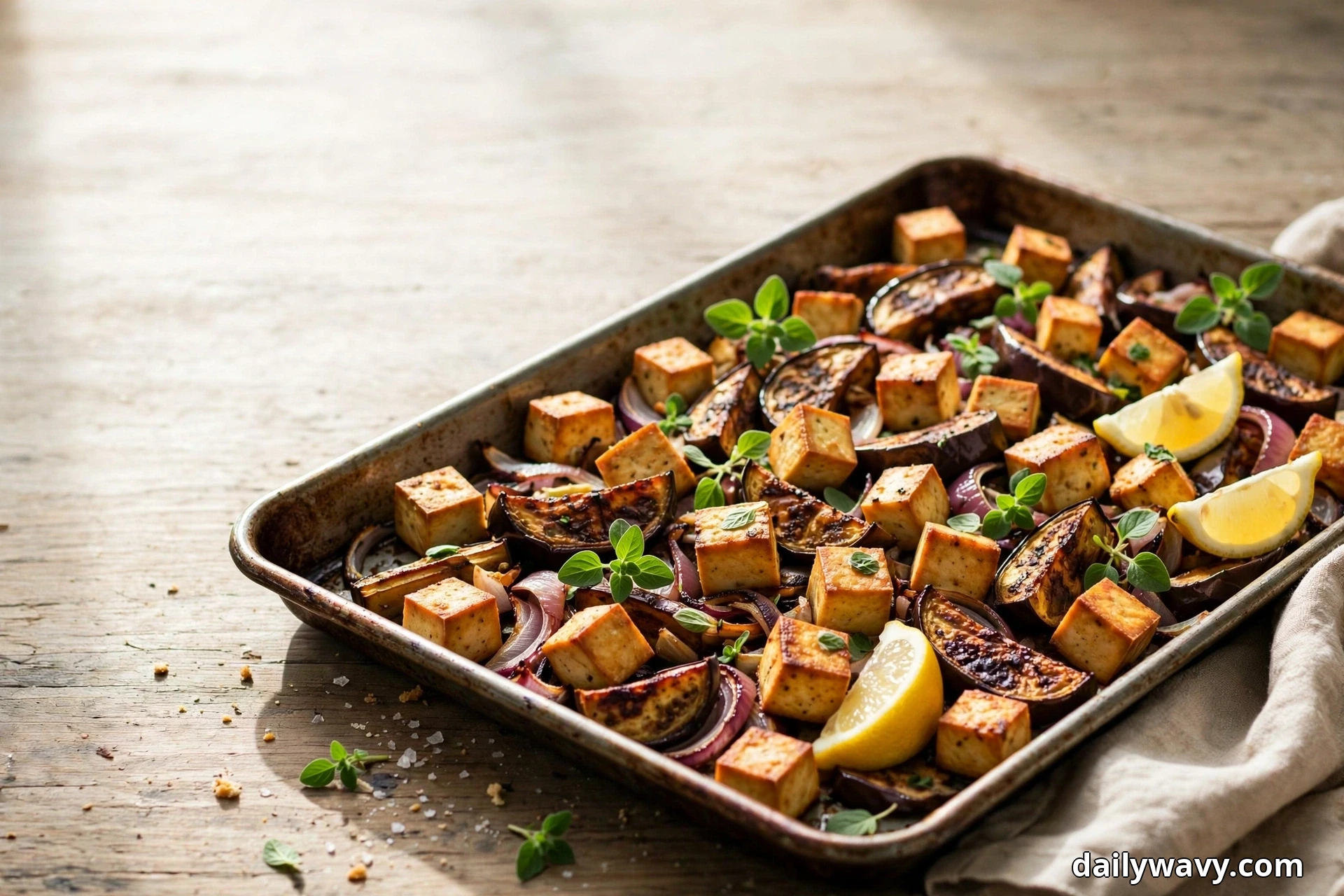 A metal sheet pan of golden roasted tofu cubes, caramelized eggplant wedges and red onion slices topped with fresh oregano leaves and lemon wedges sits on a rustic wooden table