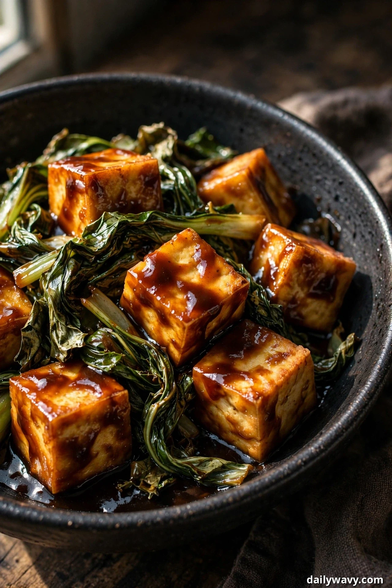Golden tofu cubes and wilted leafy greens are coated in a glossy dark hoisin sauce in a black bowl.