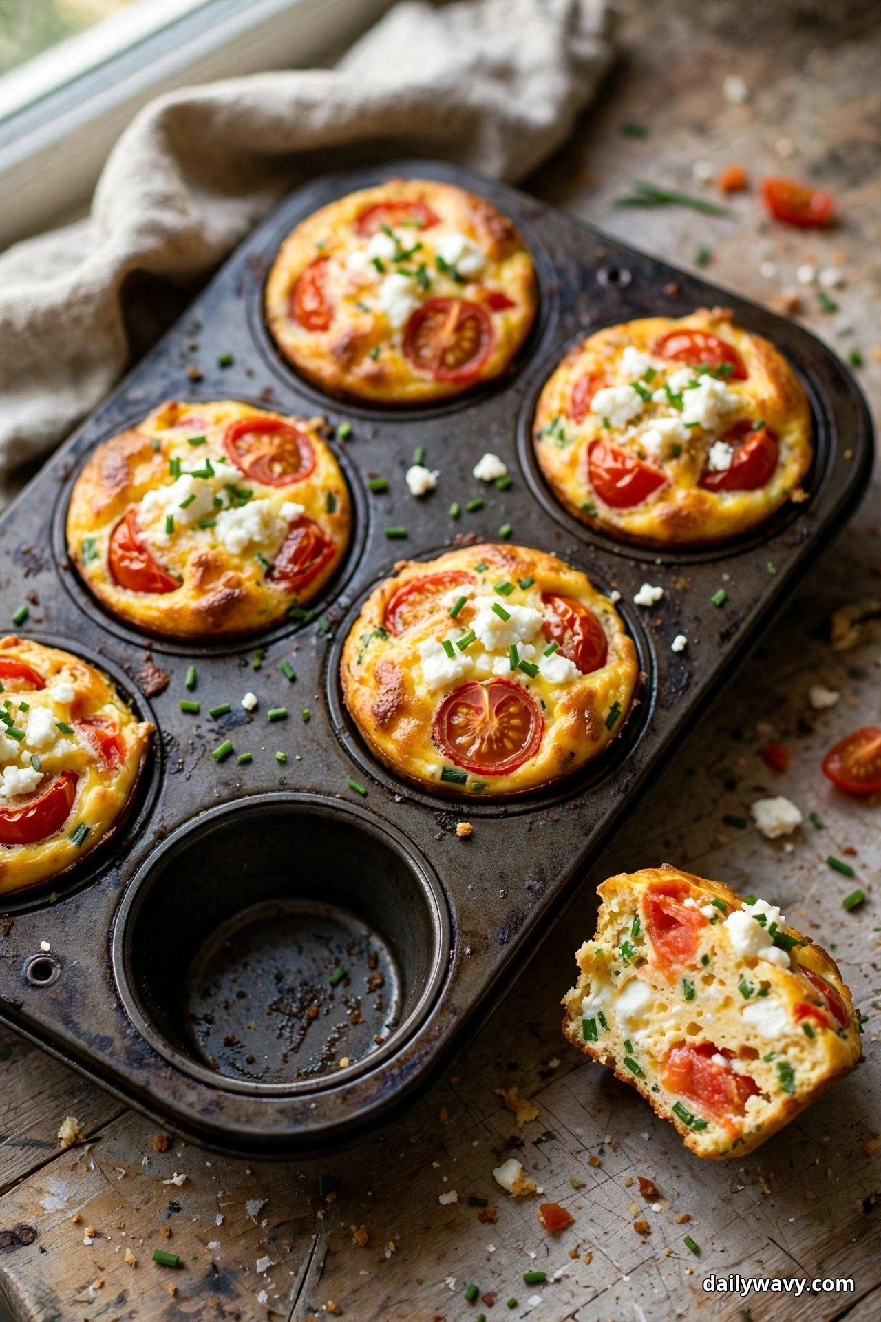 Tomato egg muffins topped with crumbled goat cheese and snipped chives sit in a muffin tray with one cup empty, with one muffin cut open beside the tray showing the cheese pockets and tomato pieces inside