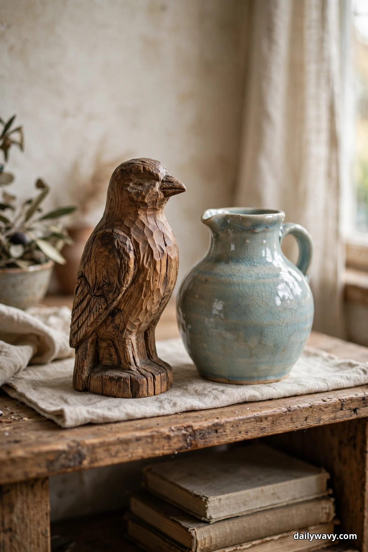 A hand carved wooden bird figure stands on a linen cloth beside a crackle glazed pale blue ceramic pitcher on a rustic wood table, with a stack of old books on the shelf below and a curtained window at the right