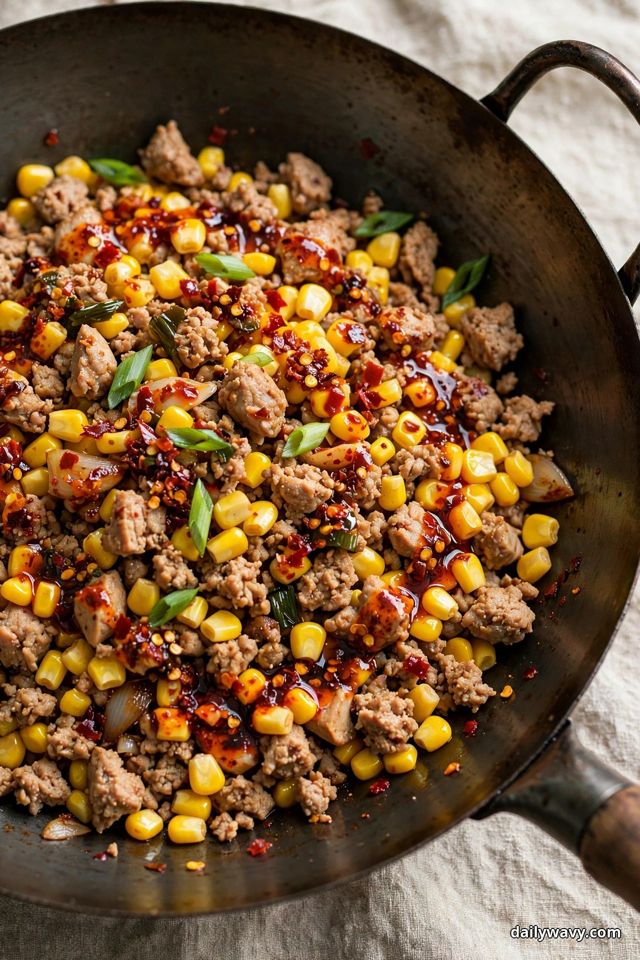 A close overhead view of a dark wok holds ground turkey crumbles and corn kernels glossed with red chili crisp oil and finished with sliced scallion greens