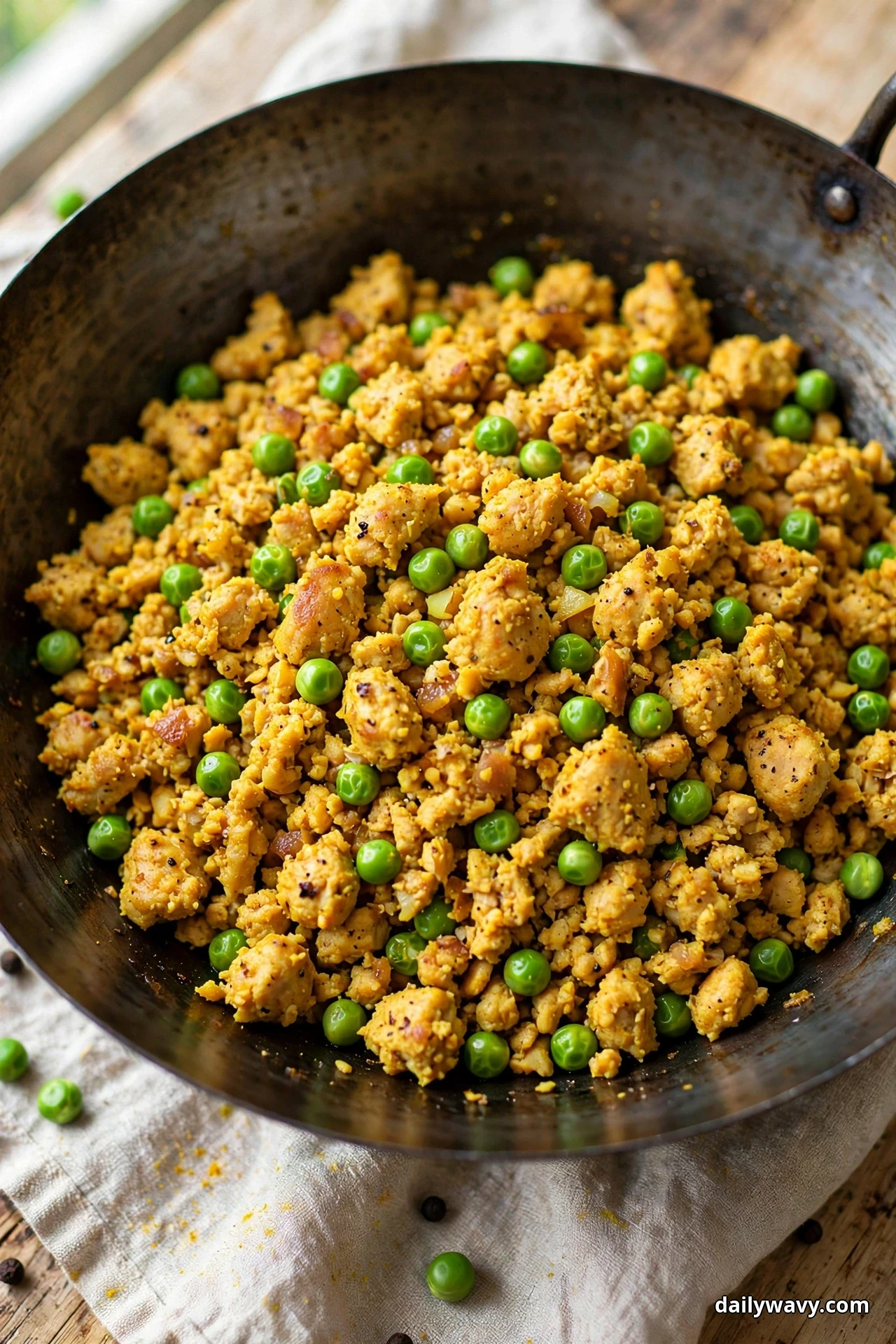 A dark wok holds golden turmeric-stained ground turkey crumbles mixed with bright green peas over a linen cloth with loose peas scattered beside it