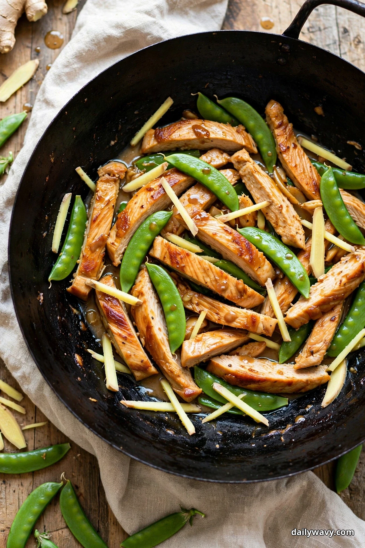A dark wok holds glossy sliced turkey strips with bright green snap peas and pale ginger matchsticks on a linen cloth