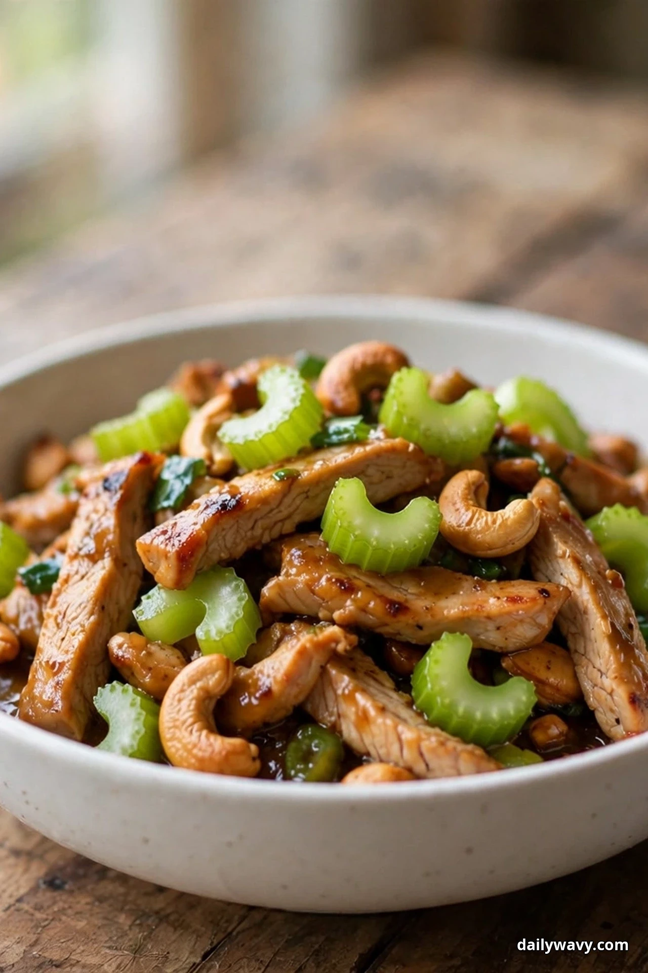 A white bowl of sliced glazed turkey strips is mixed with bright green celery slices and whole roasted cashews on a wooden table