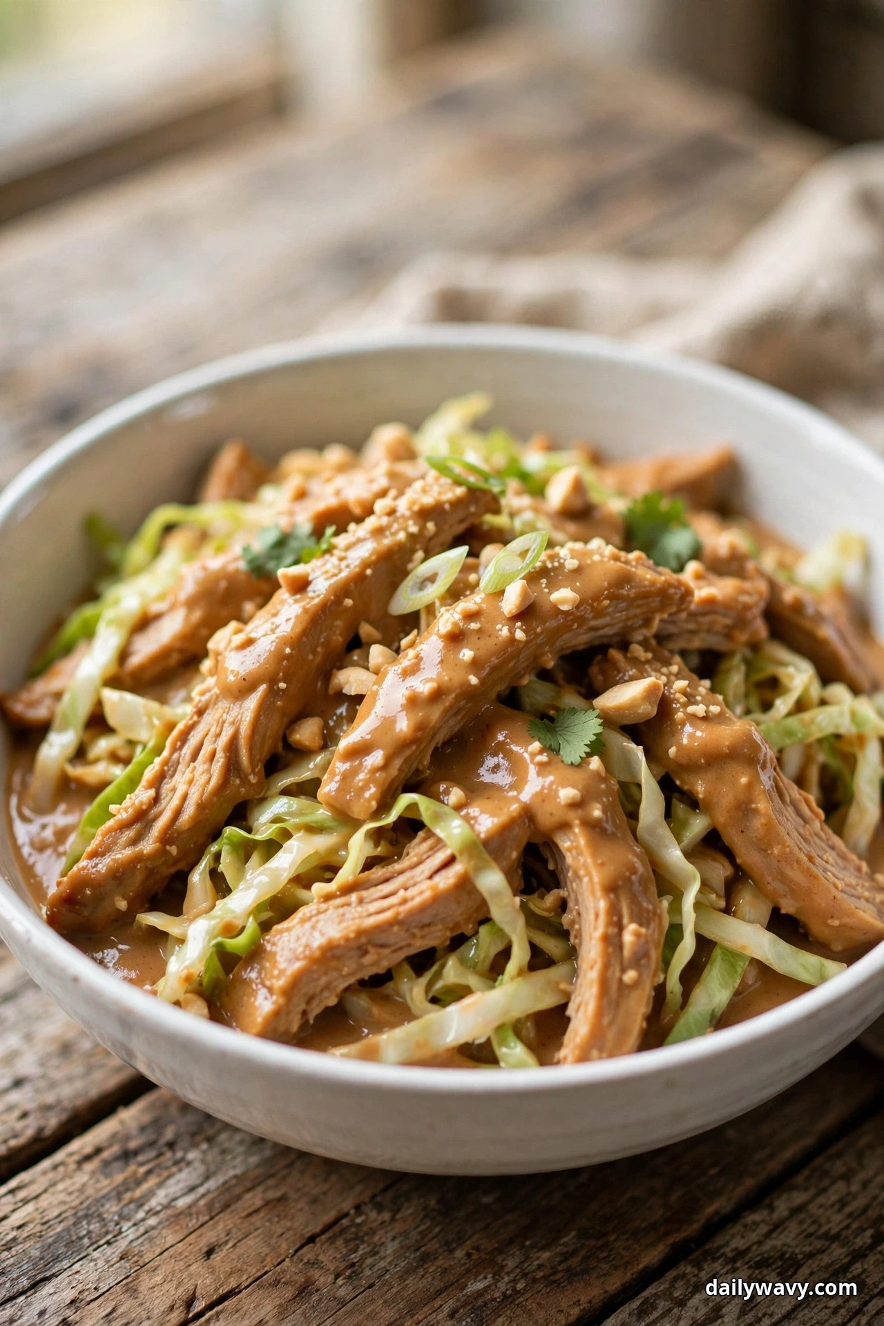 A white bowl of sliced turkey strips coated in peanut sauce rests over shredded green cabbage and is topped with crushed peanuts and cilantro leaves
