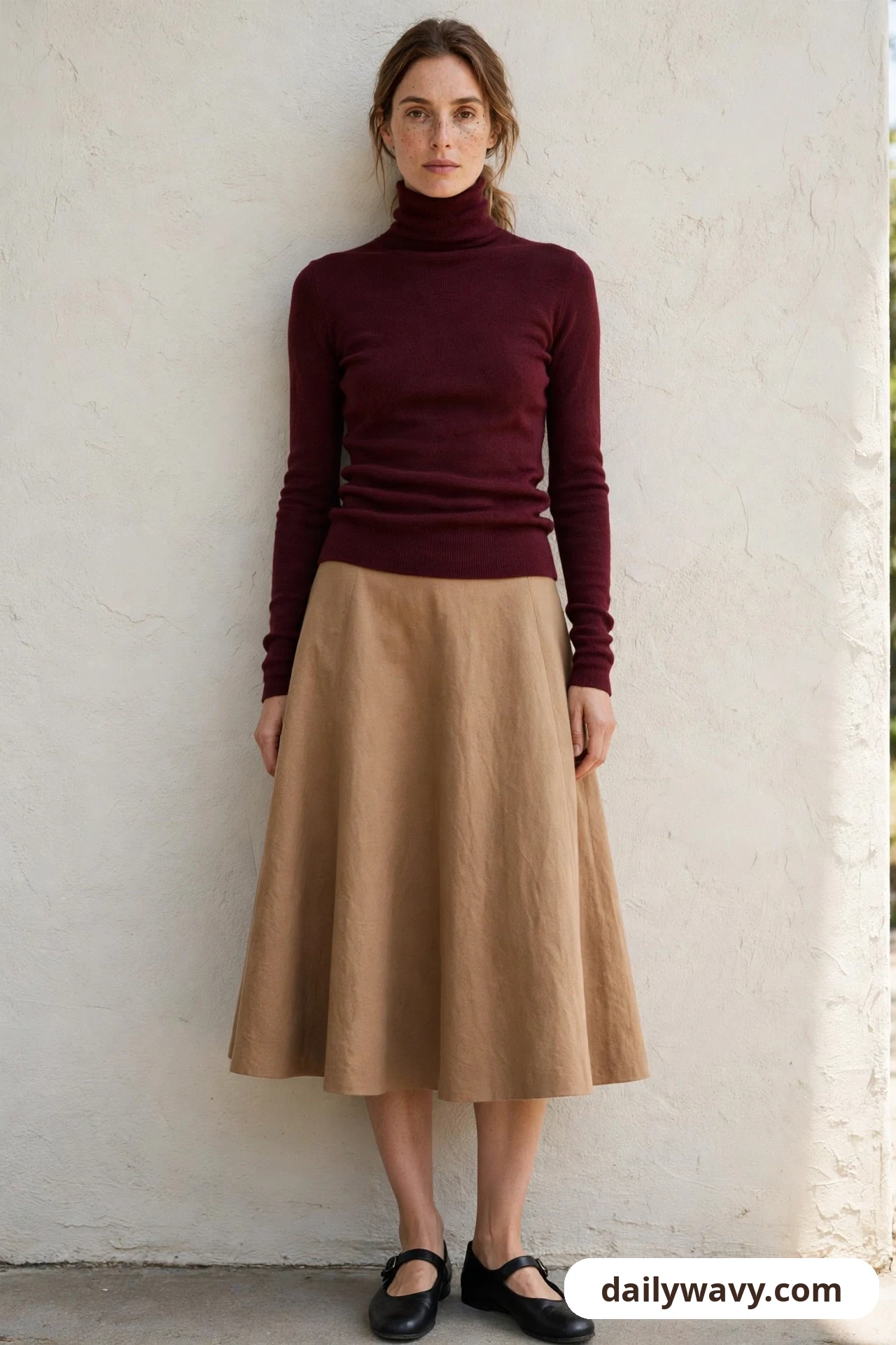 A woman stands against a cream wall wearing a burgundy fitted turtleneck with a camel A-line midi skirt and black Mary Jane flats.