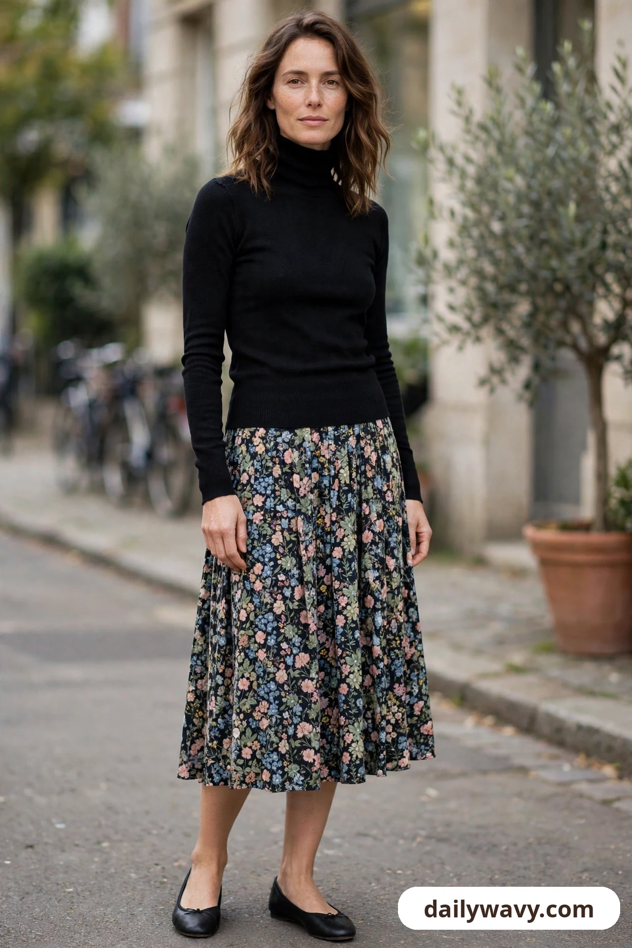 A woman stands on a street wearing a black fitted turtleneck with a floral printed midi skirt and black ballet flats.