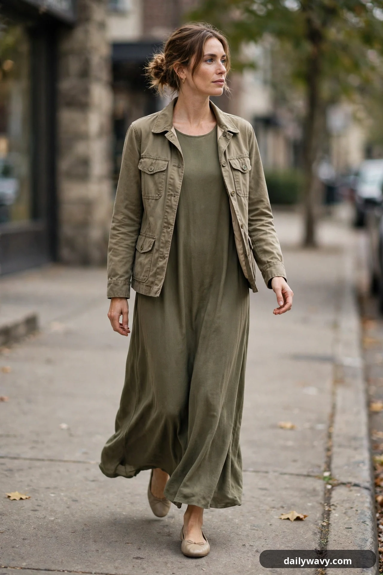 A woman wears a khaki utility jacket open over an olive maxi dress with neutral ballet flats.