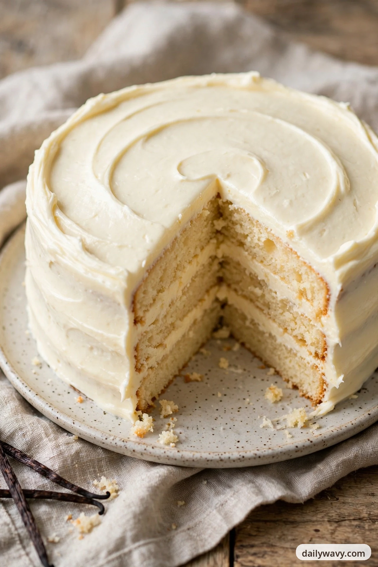 A three-layer vanilla cake covered in smooth white mascarpone frosting swirled across the top sits on a speckled plate with one wedge removed, showing the pale layers and cream filling inside.