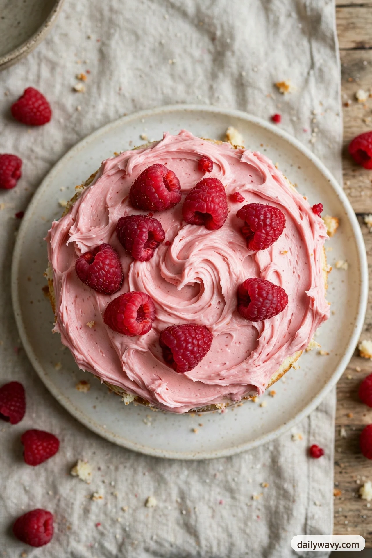 An overhead view shows a round vanilla cake on a cream plate covered in swirled pink raspberry buttercream and topped with fresh raspberries, with more raspberries scattered on the linen and wooden table around the plate.