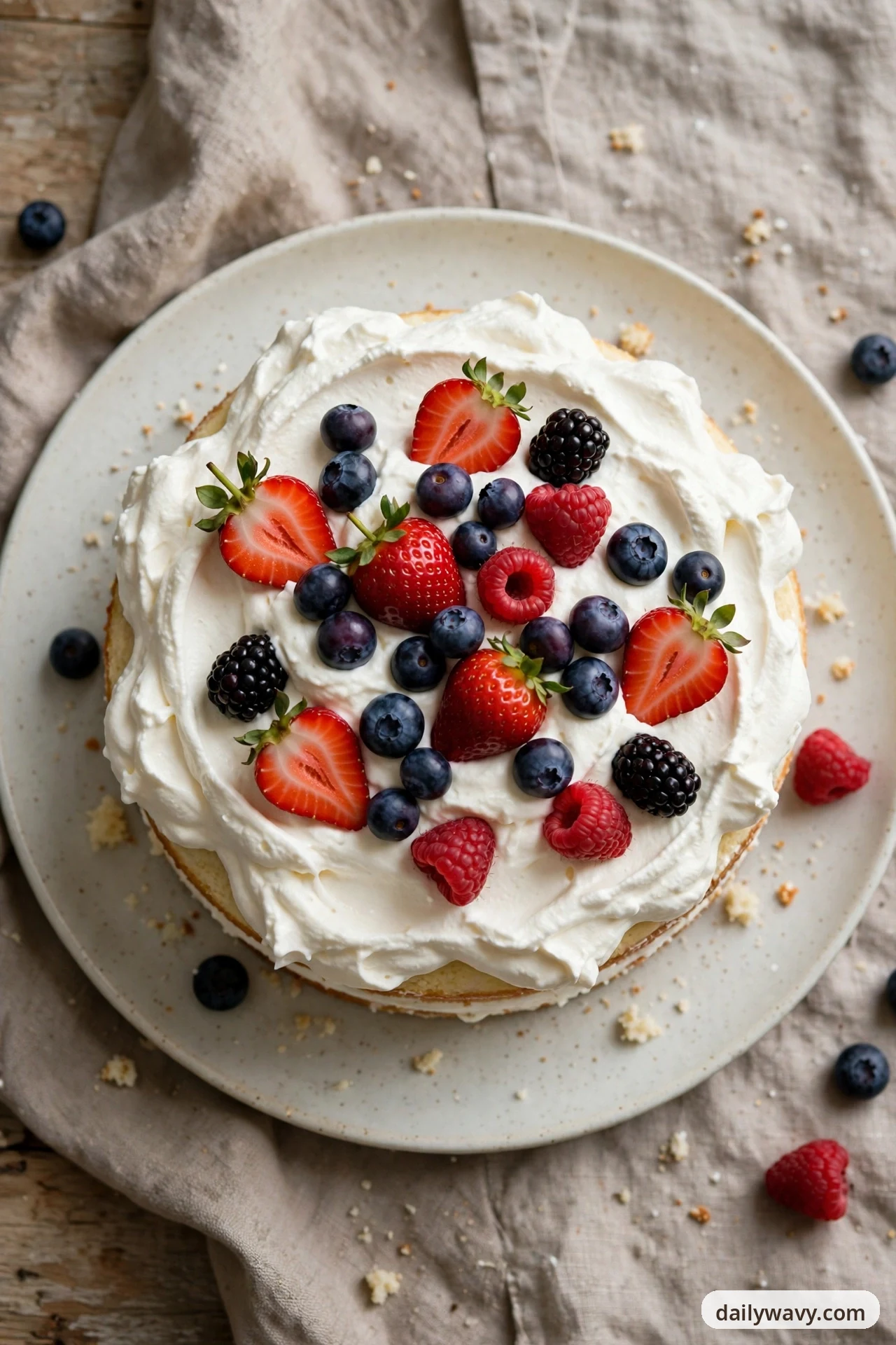 An overhead view shows a round vanilla cake on a cream plate covered in billowy whipped cream and topped with halved strawberries, blueberries, raspberries and blackberries, with a few loose berries scattered on the plate and linen around it.