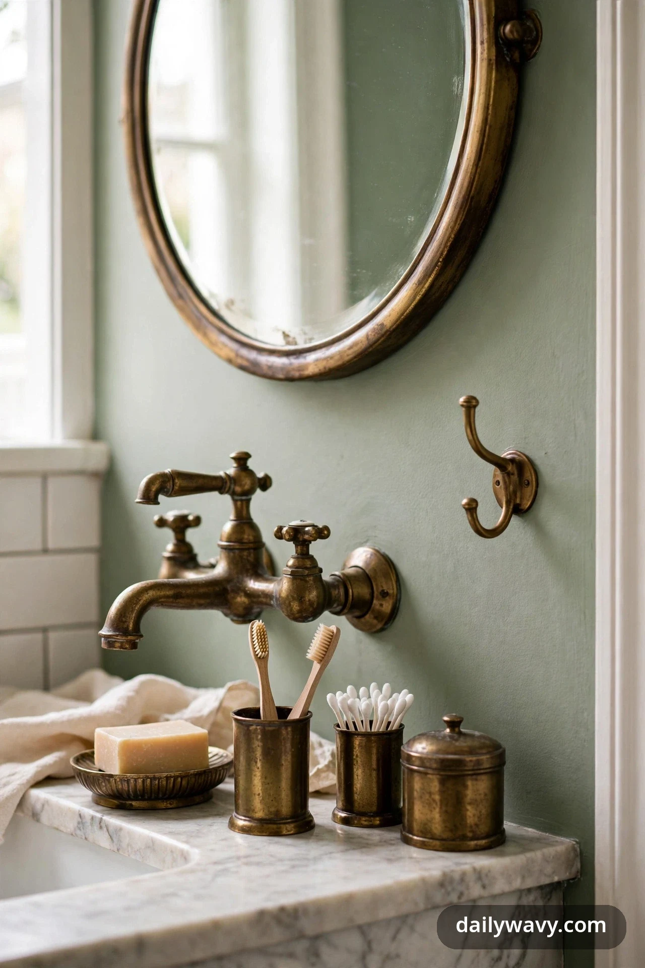 Aged brass fixtures repeat across a bathroom wall and marble counter, in a wall-mounted faucet, a double hook, an oval mirror frame, a soap dish, and lidded canisters holding toothbrushes and cotton swabs.