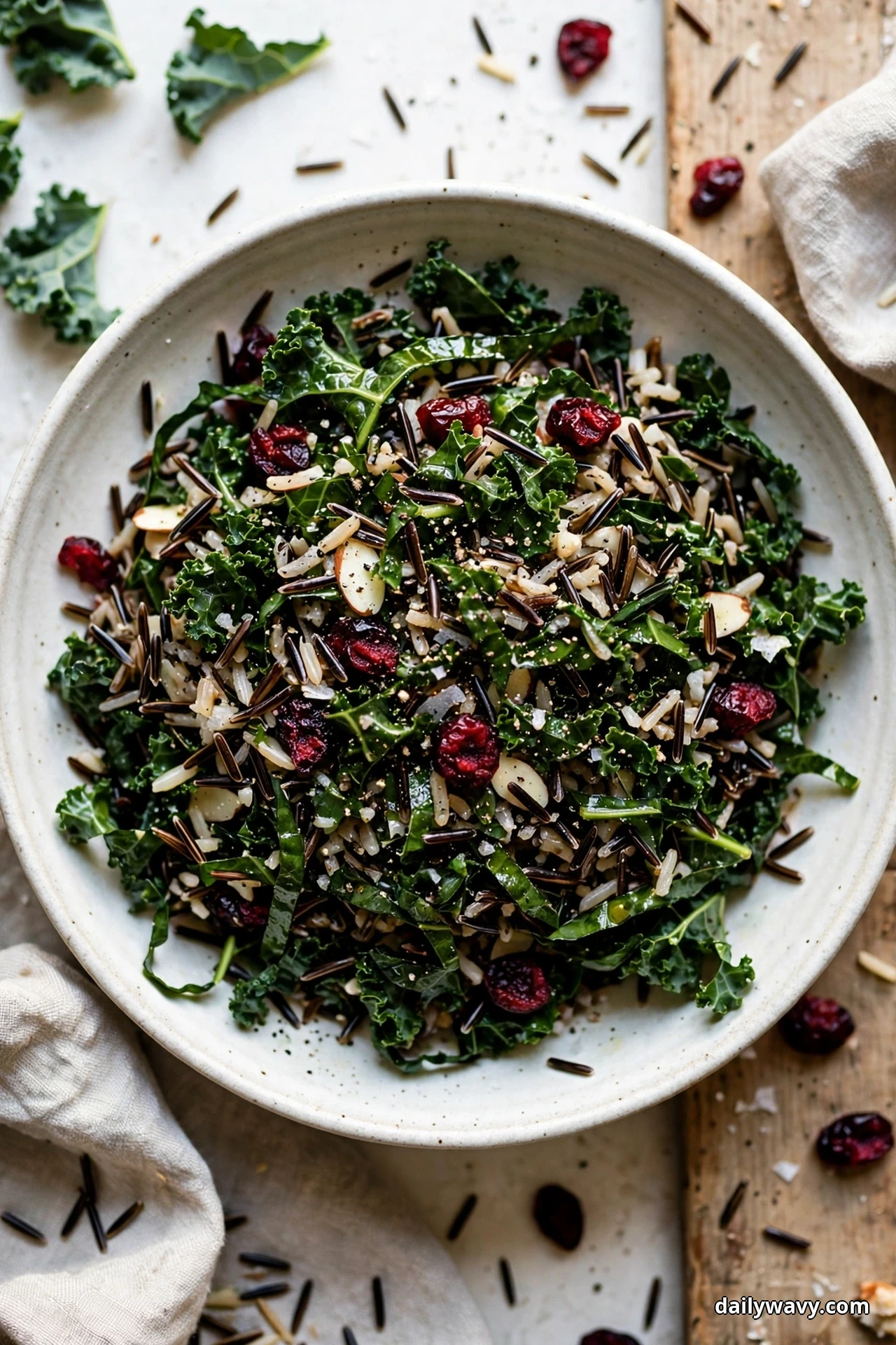 An overhead view shows a bowl of wild rice salad with massaged kale ribbons and dried cranberries scattered over the top