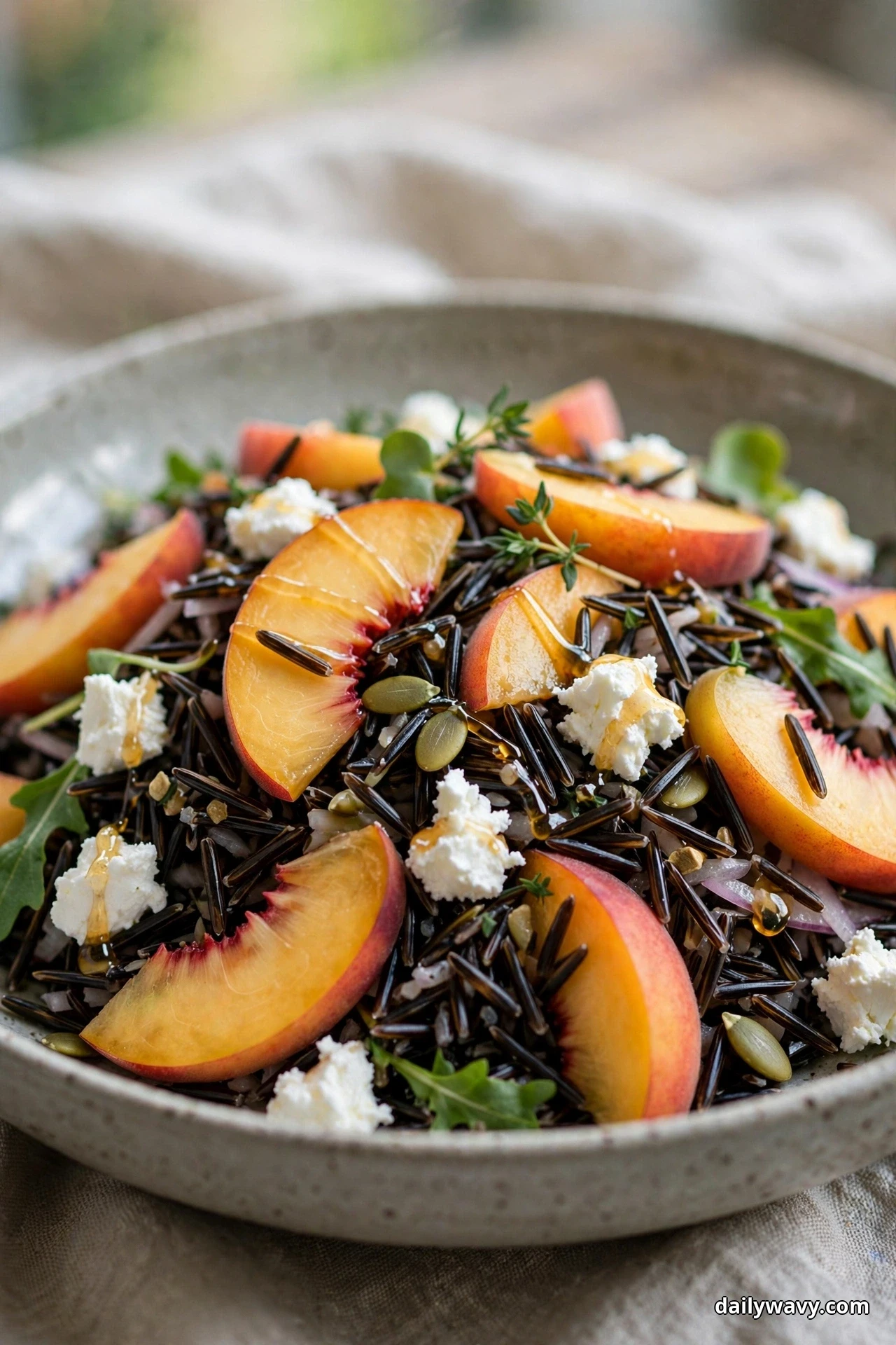 A bowl of wild rice salad is topped with fresh peach slices and crumbled goat cheese with a honey drizzle